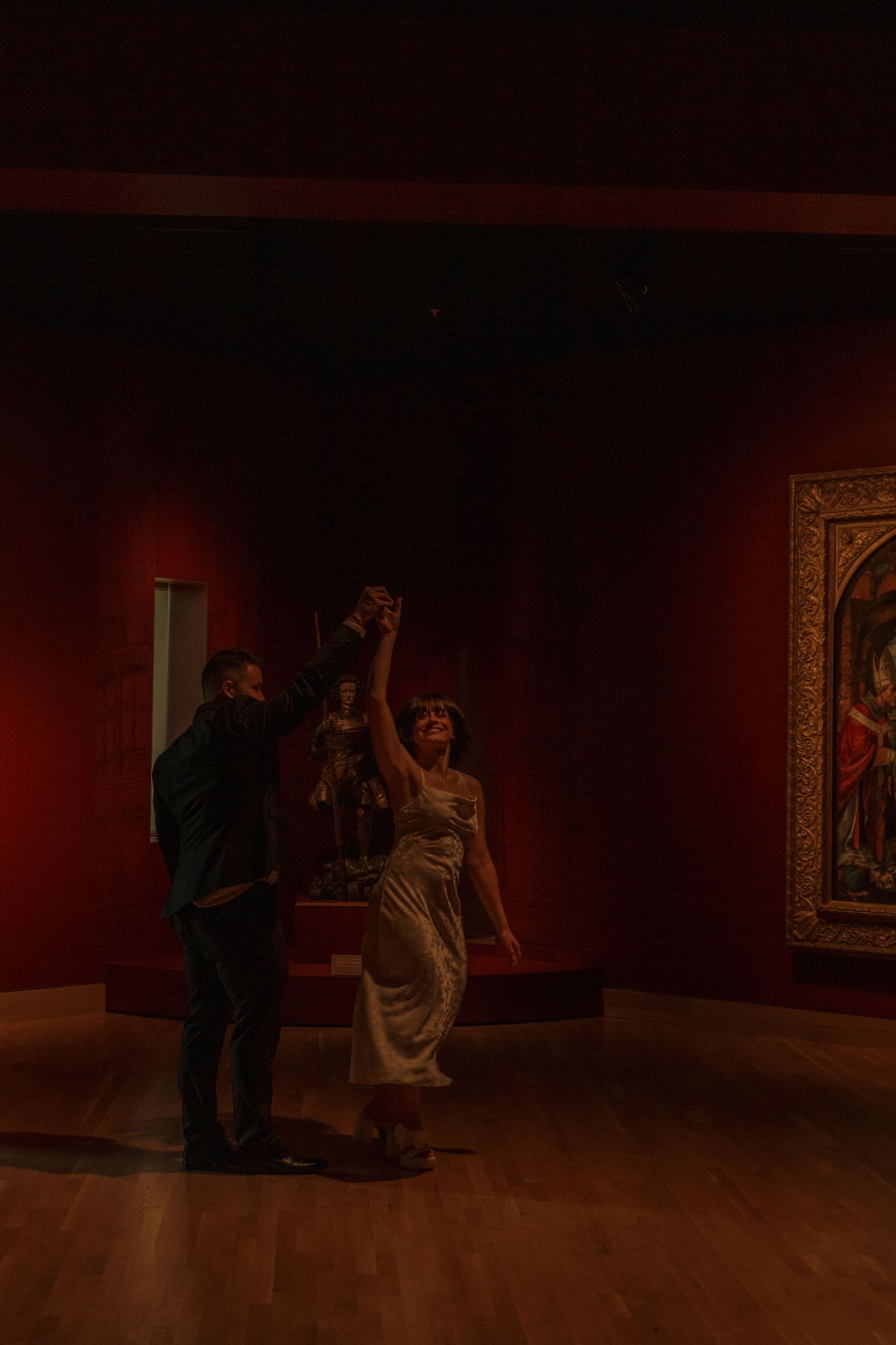 Two figures view artwork on dark red walls in a dimly lit museum gallery space.