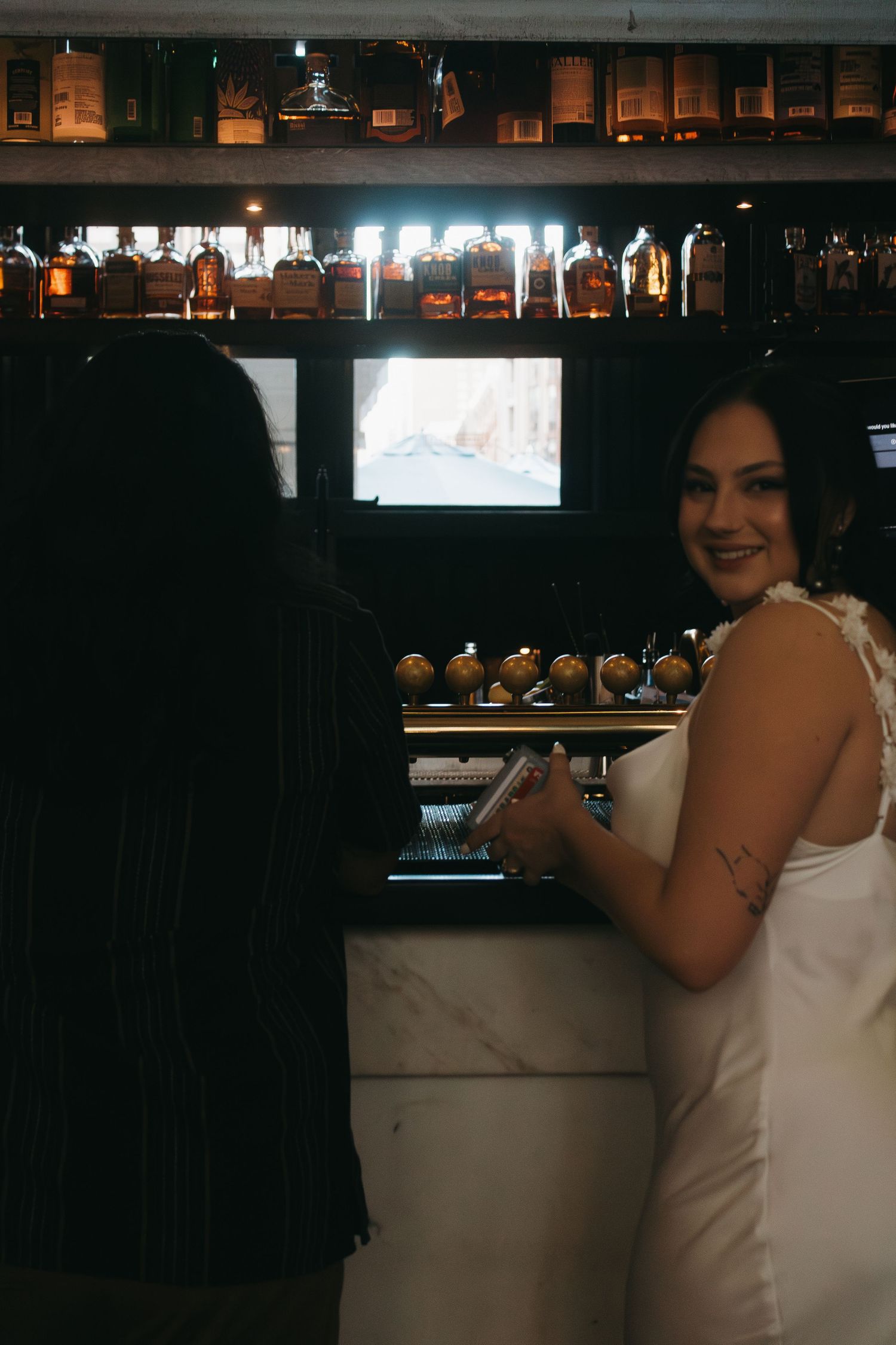 A bartender stands behind a dimly lit bar with bottles and glassware on display.