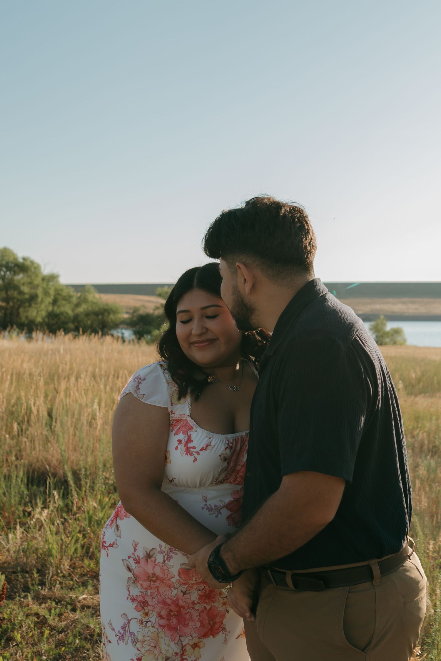 A couple shares a sweet embrace against a backdrop of rolling hills and a serene lake.
