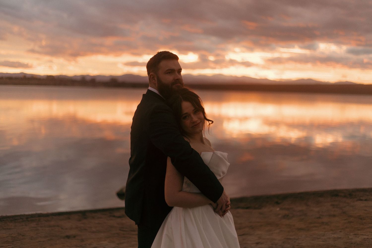 Wedding couple embracing on a beach at sunset with golden clouds in the background.