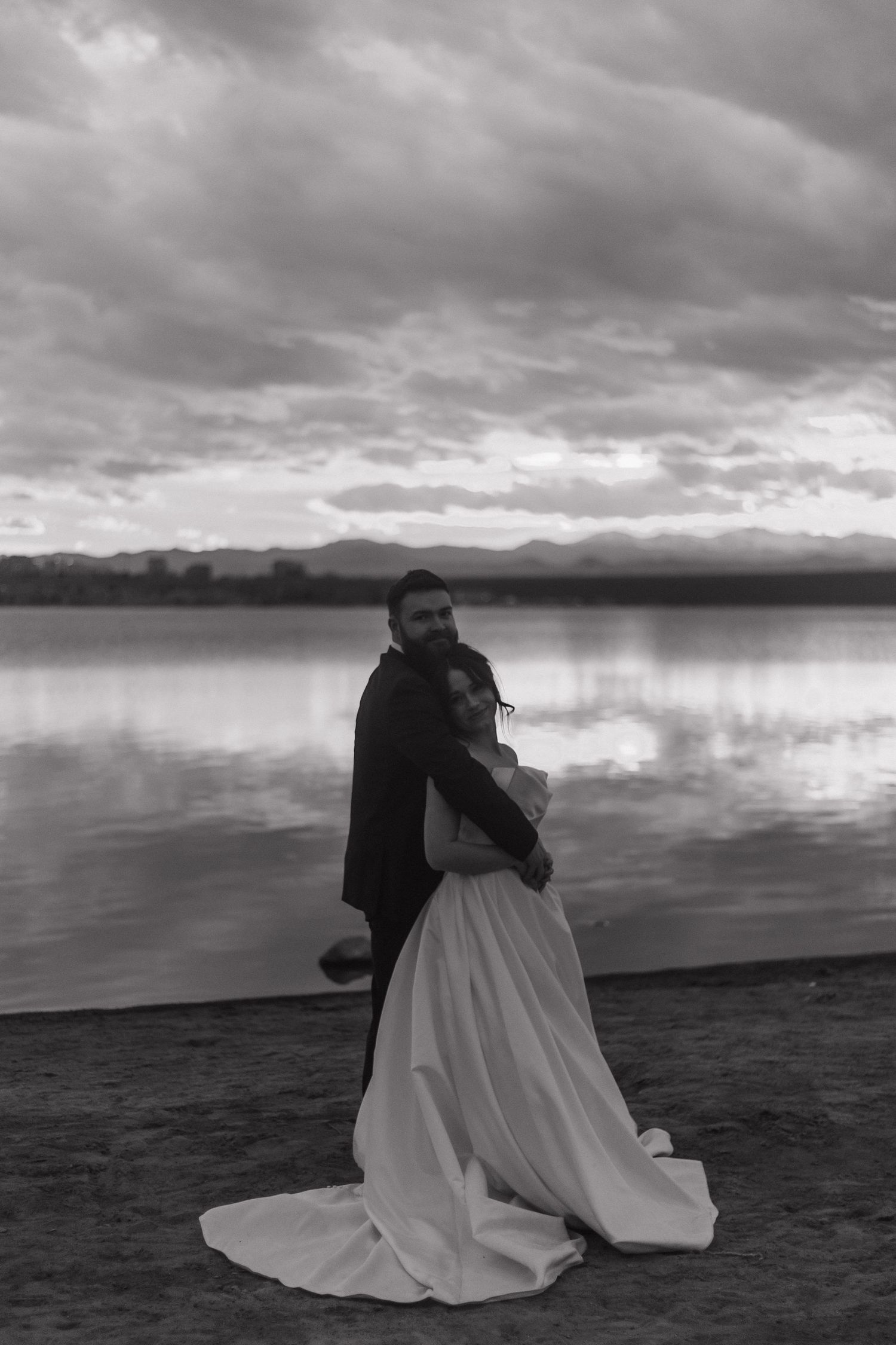 A black and white wedding photo captures a couple by a scenic lake with dramatic clouds overhead.