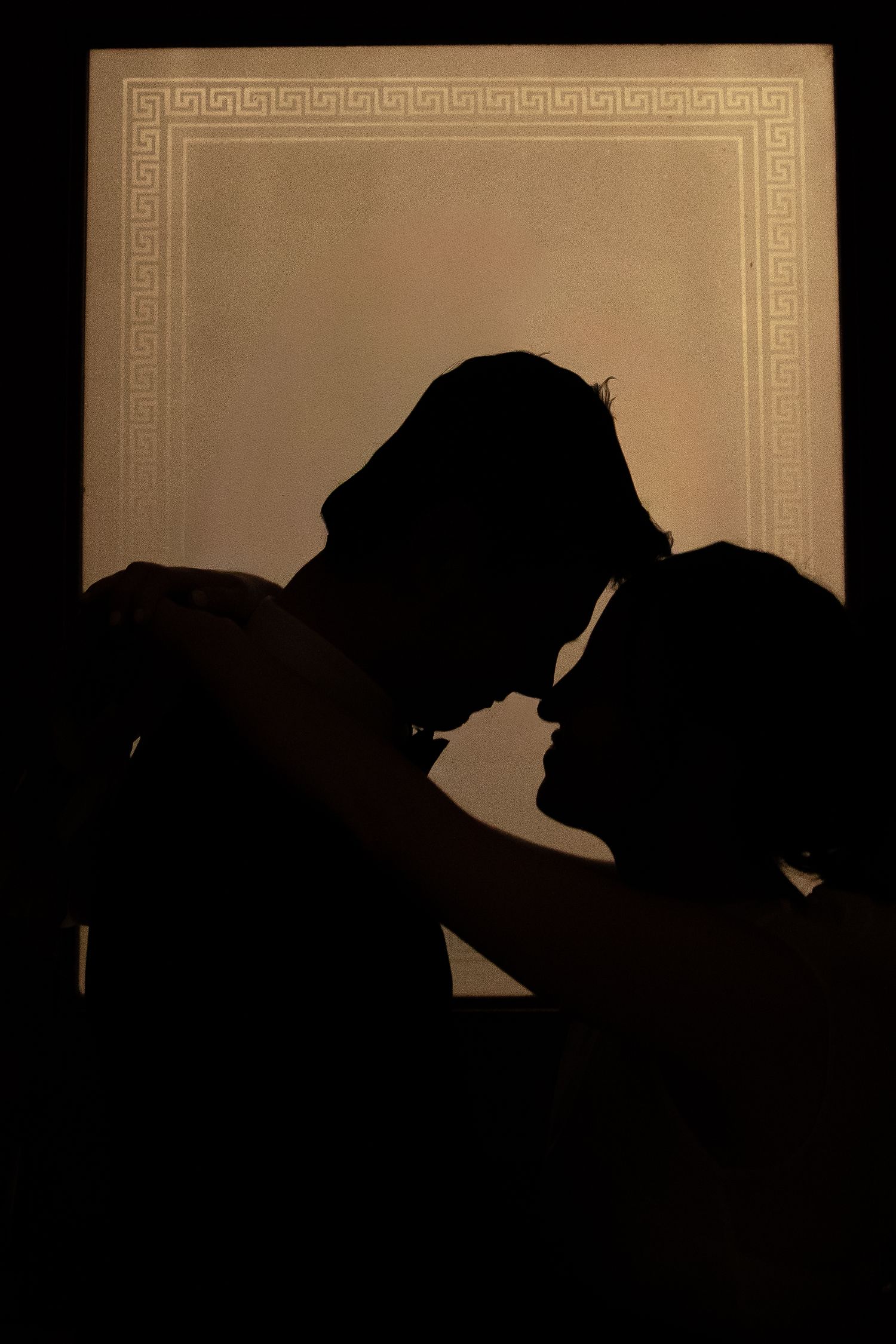 Artistic silhouette of couple against backlit wall with Greek key pattern border.
