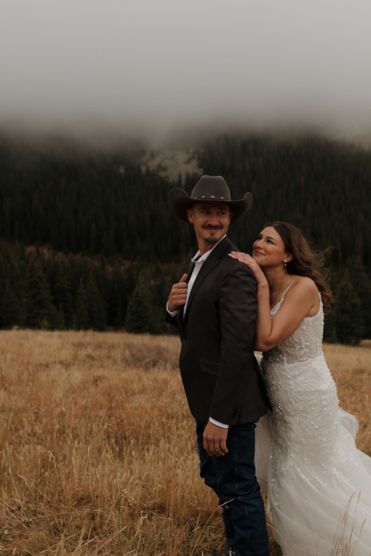 A joyful wedding couple poses in a mountain field at dusk.
