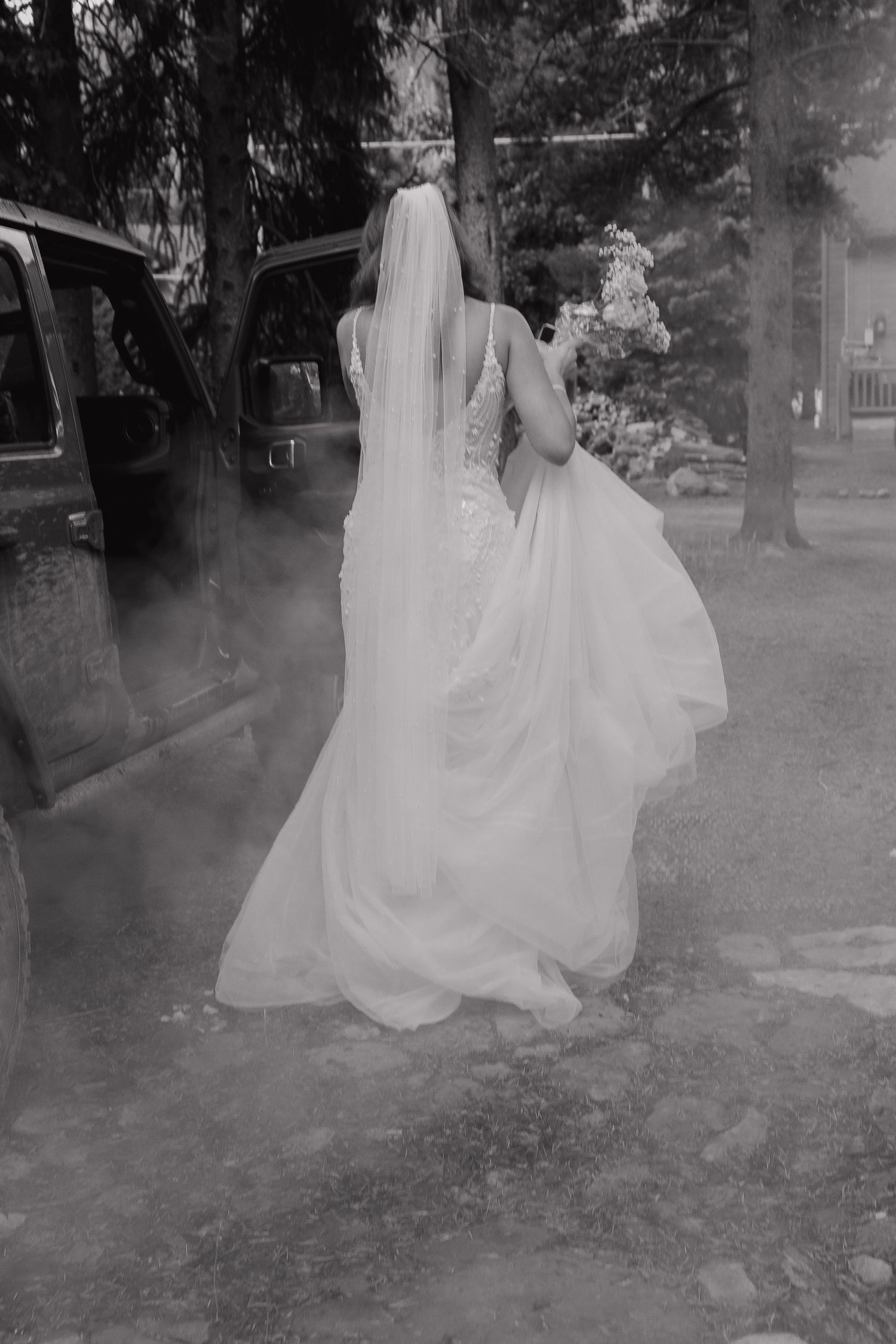 A bride in a flowing white dress and veil stands near a car door in a misty black and white photograph.