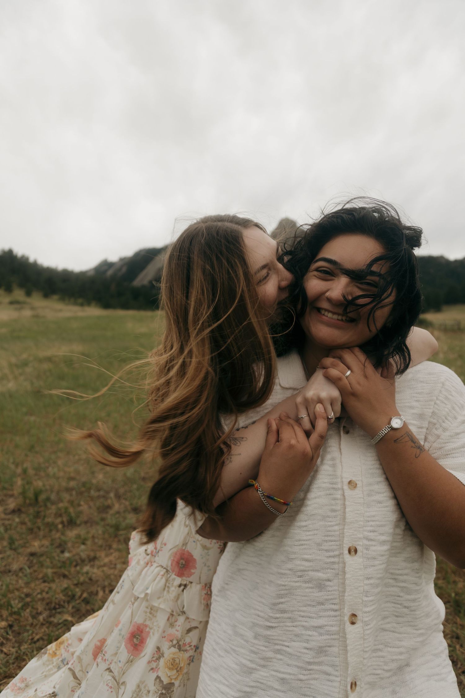 Two women embrace and laugh in a green meadow with rocky mountains in the background on a cloudy day.