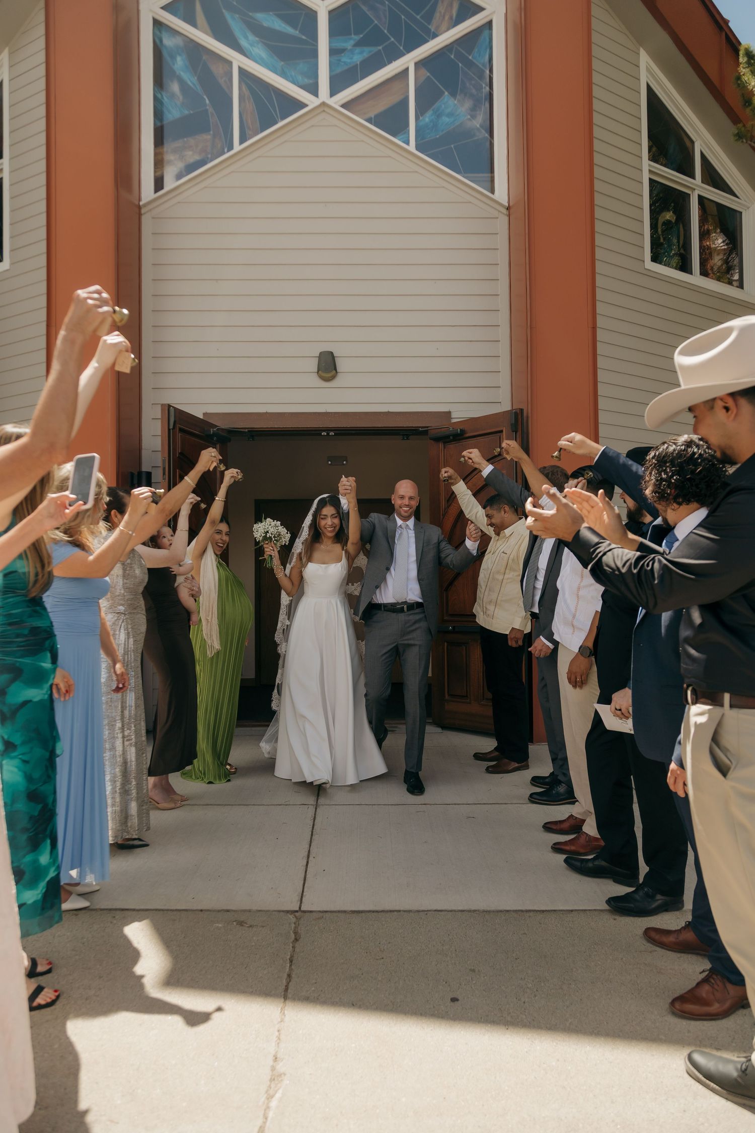 Newlyweds exit church triumphantly as guests raise hands in celebration during outdoor wedding send-off.