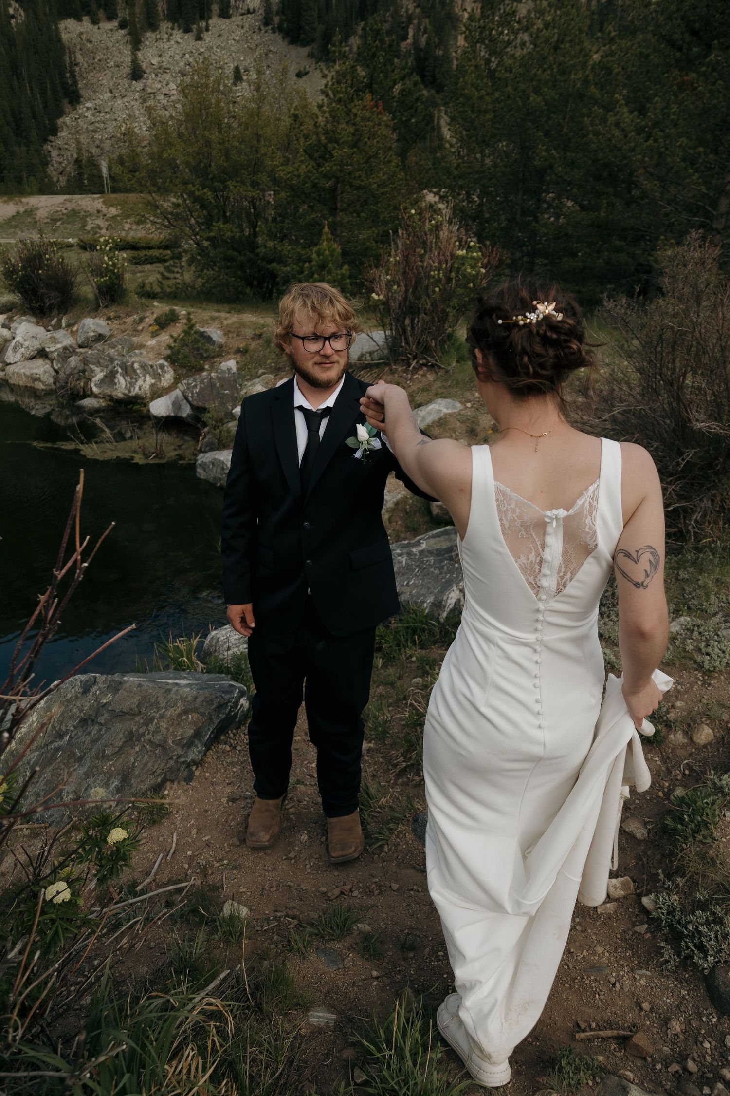 Bride in lace-back white gown adjusts groom's boutonniere near scenic mountain lake setting.