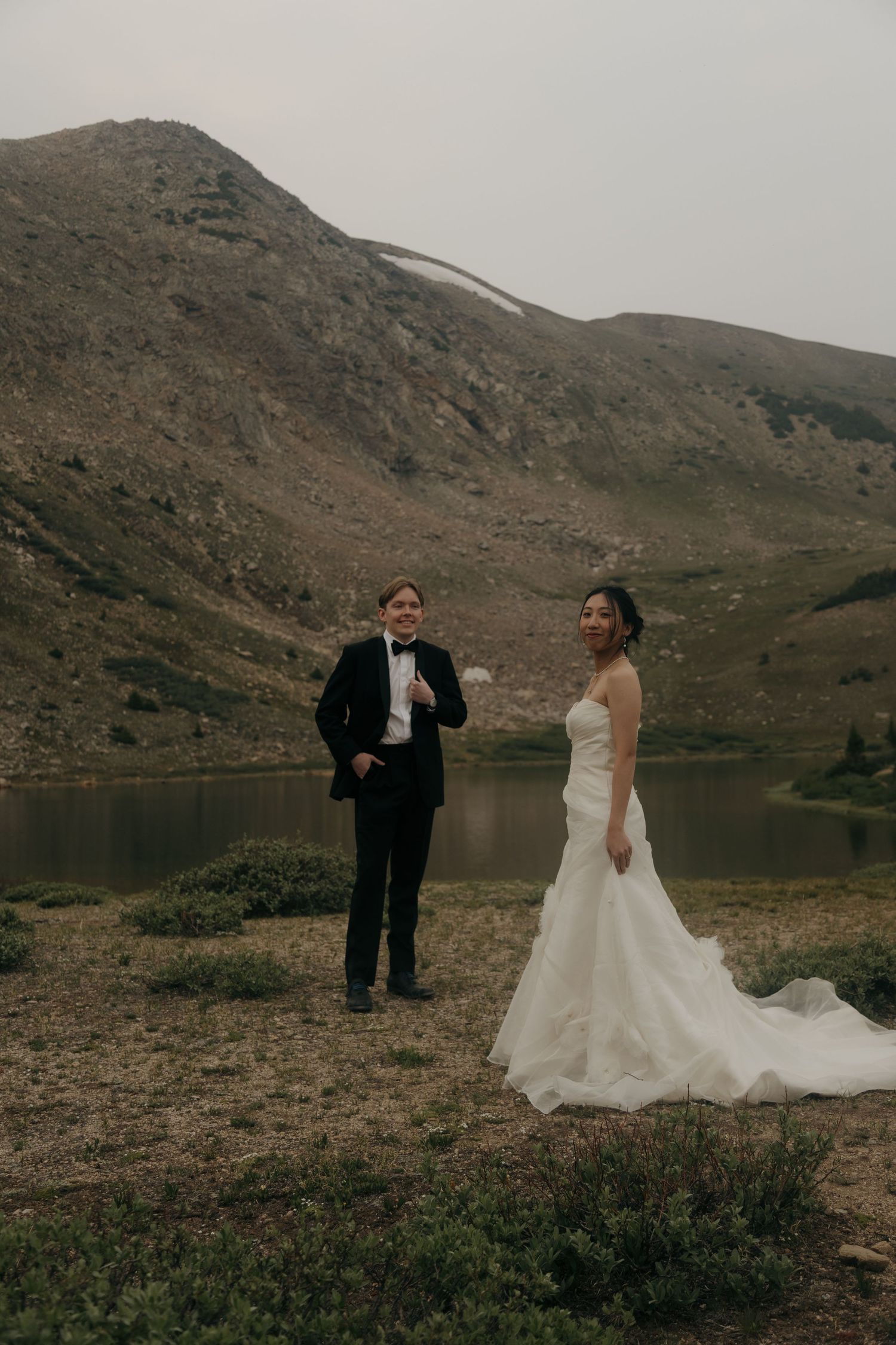 Wedding couple poses before a misty alpine lake, bride in flowing white gown and groom in classic black tuxedo.