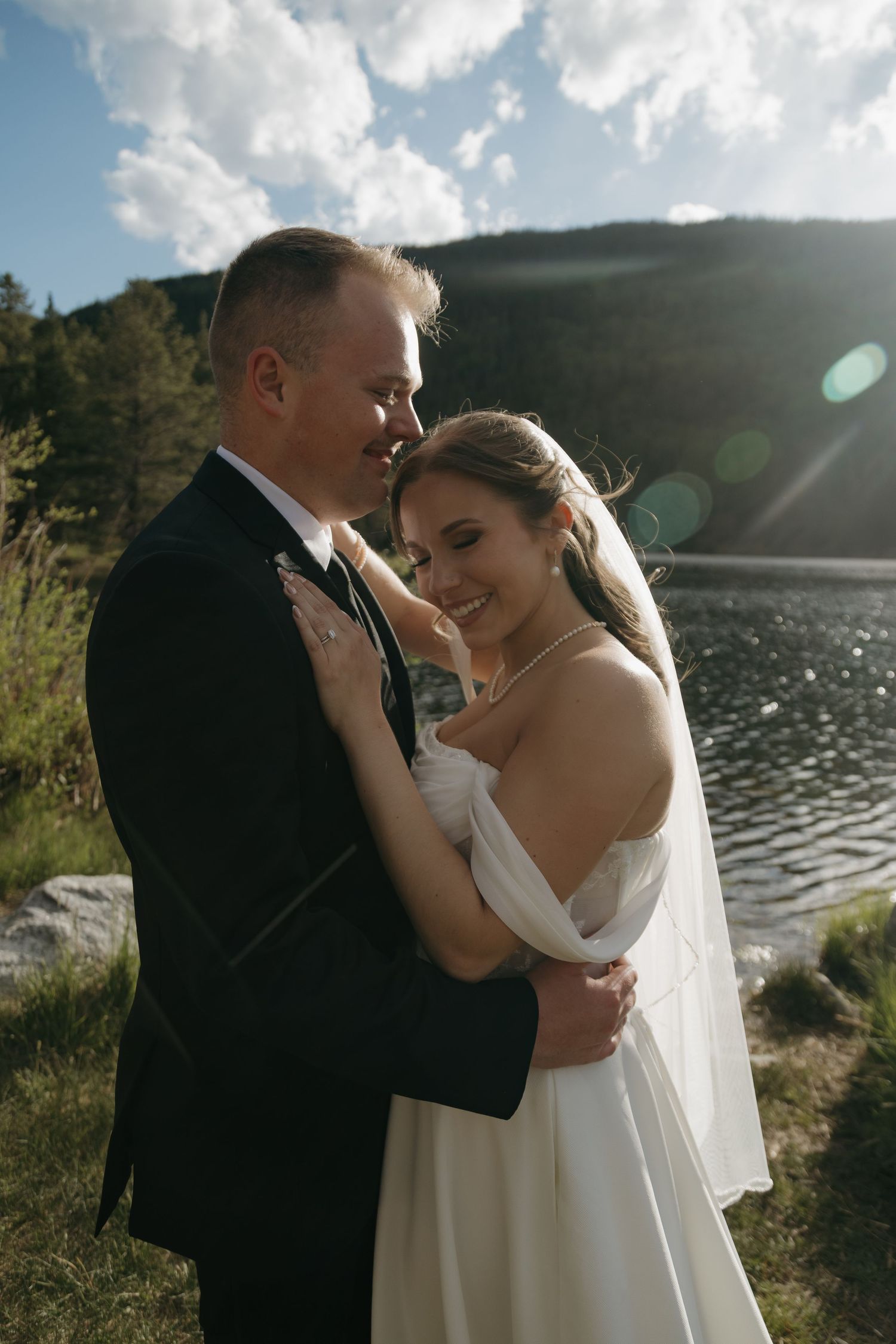 Joyful bride and groom embrace near a sunlit mountain lake, smiling warmly during their outdoor elopement.
