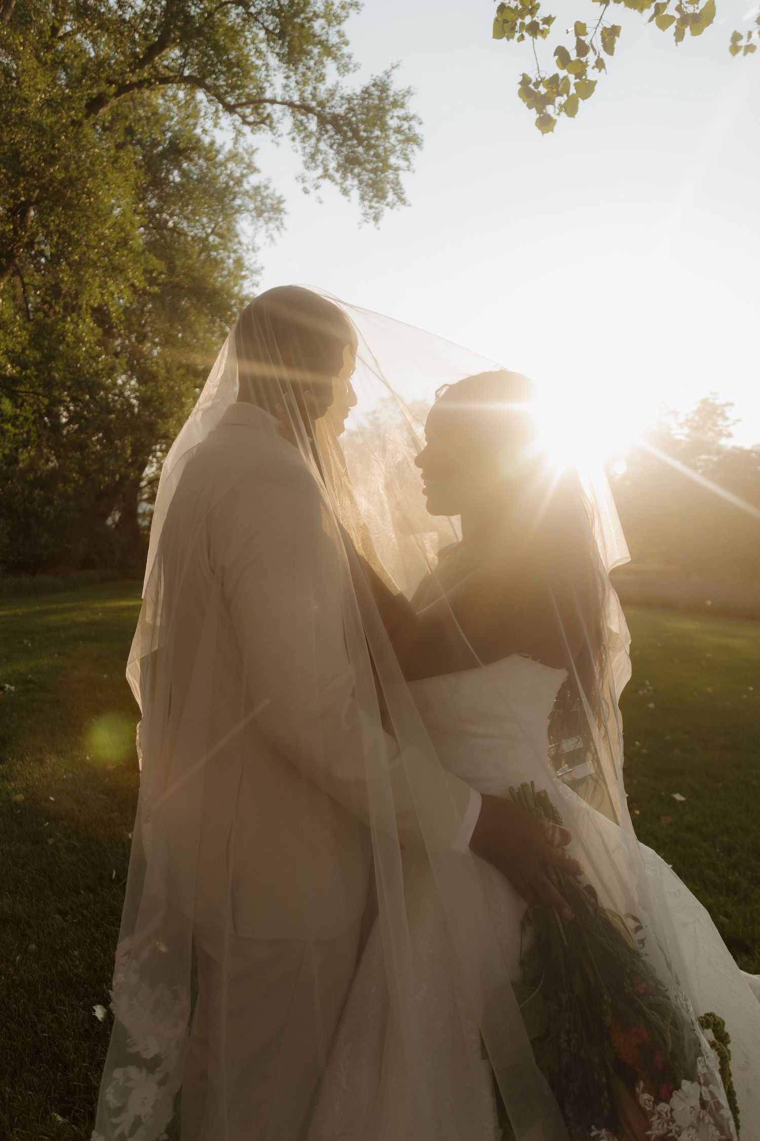Newlywed couple stands close together under a flowing veil with brilliant golden sunset light glowing behind them outdoors.
