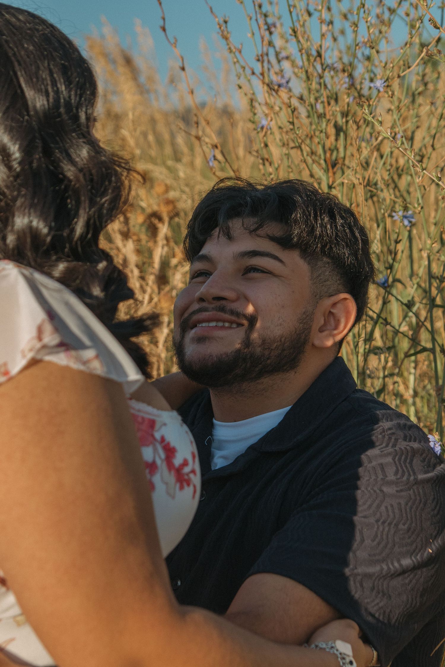 A romantic couple embraces outdoors against a backdrop of golden dried grass and blue sky.