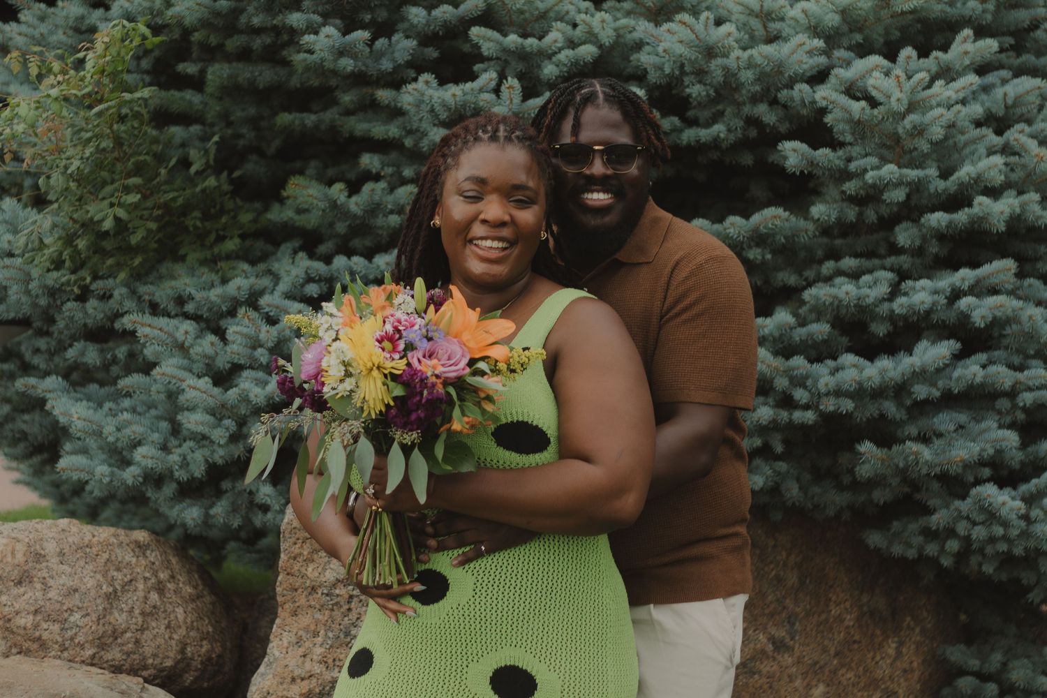 A couple surrounded by evergreen trees shares joyful moments during an outdoor engagement photo session.