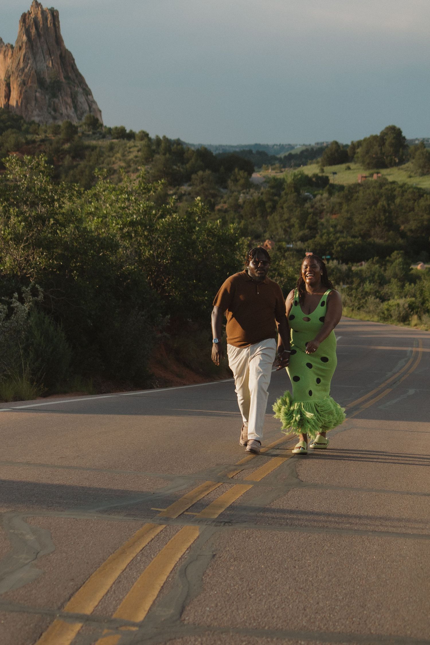 A couple walks down a scenic mountain road with dramatic rock formations in the background at sunset.