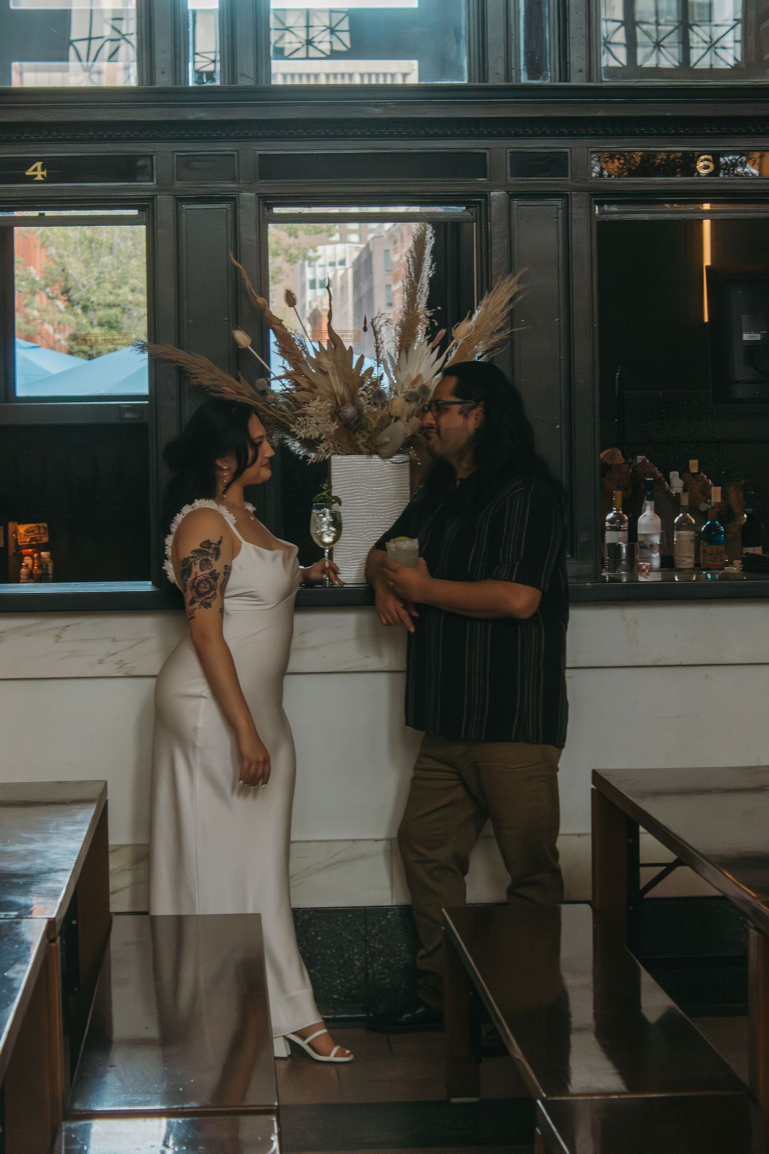 A couple in casual wedding attire stands and chats in a bar setting with pampas grass decor and liquor bottles in background.