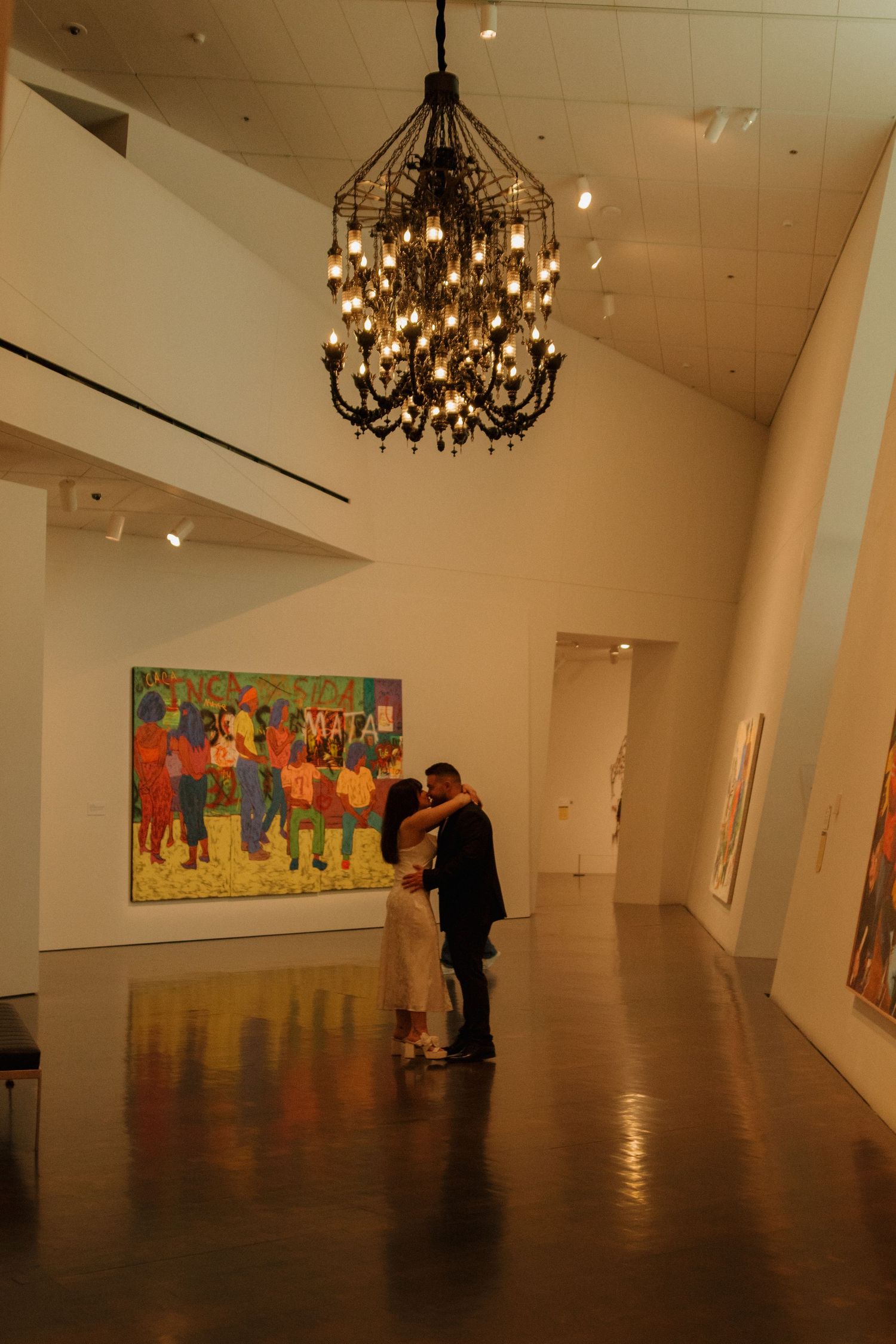 Art gallery interior with chandelier lighting and colorful paintings on display.
