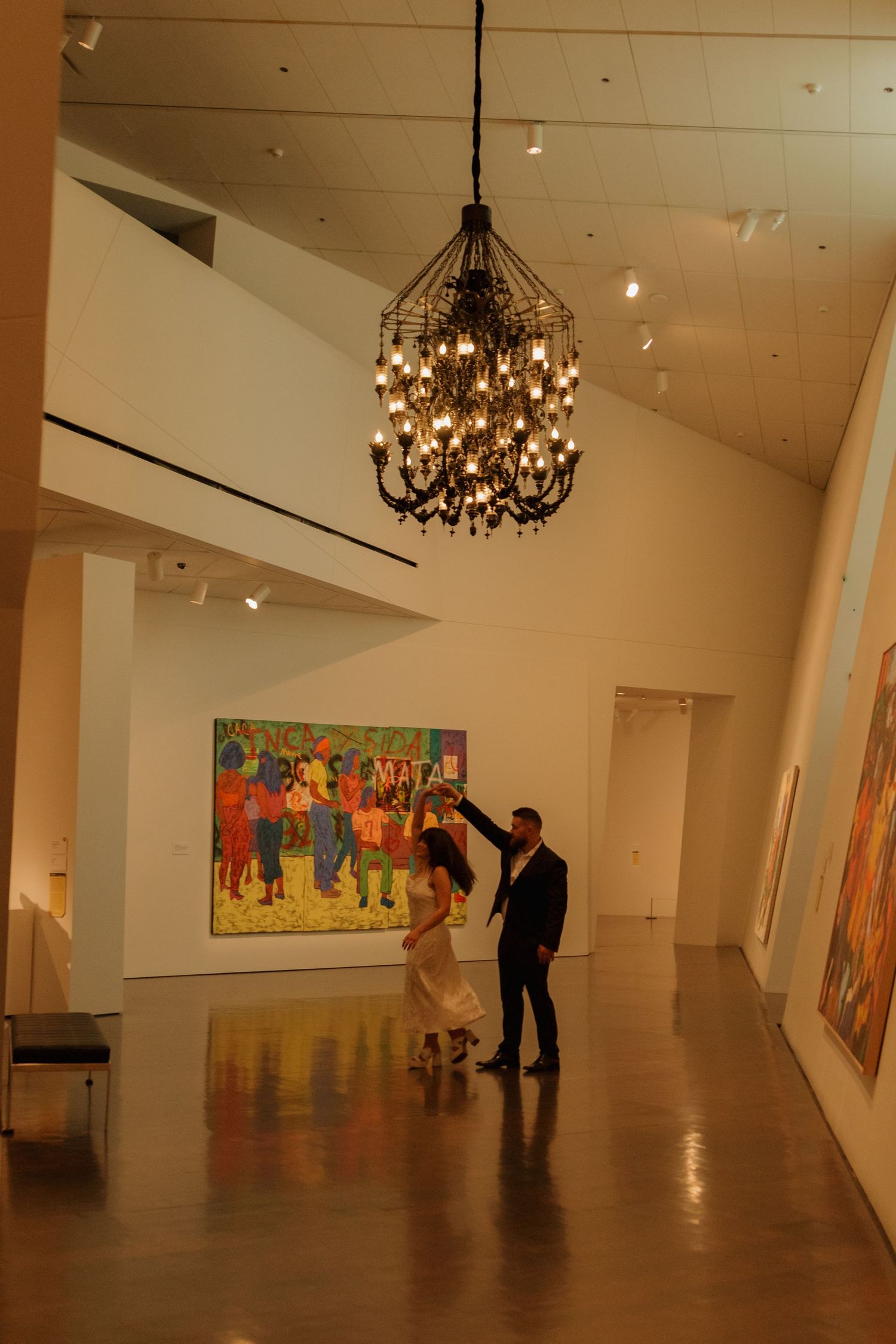 A couple views artwork in an elegant gallery space with a dramatic black chandelier hanging from the vaulted ceiling.