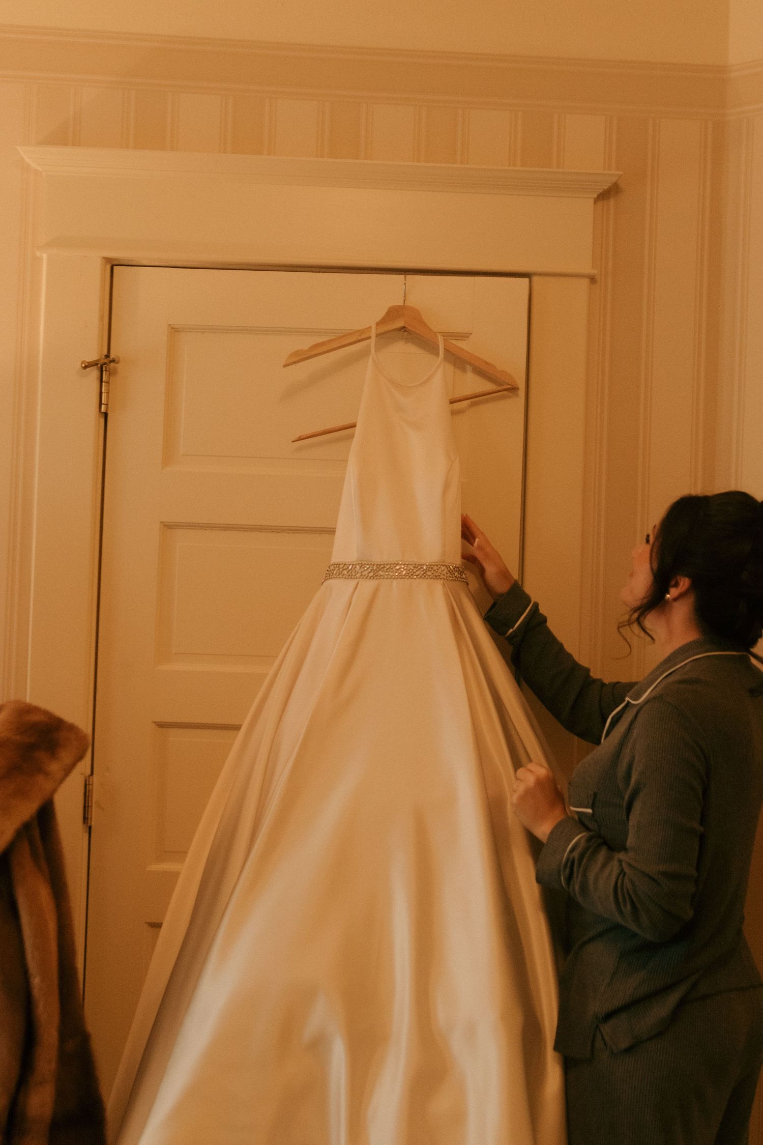 A white wedding dress hangs on a door while someone adjusts it in a warm-lit room.