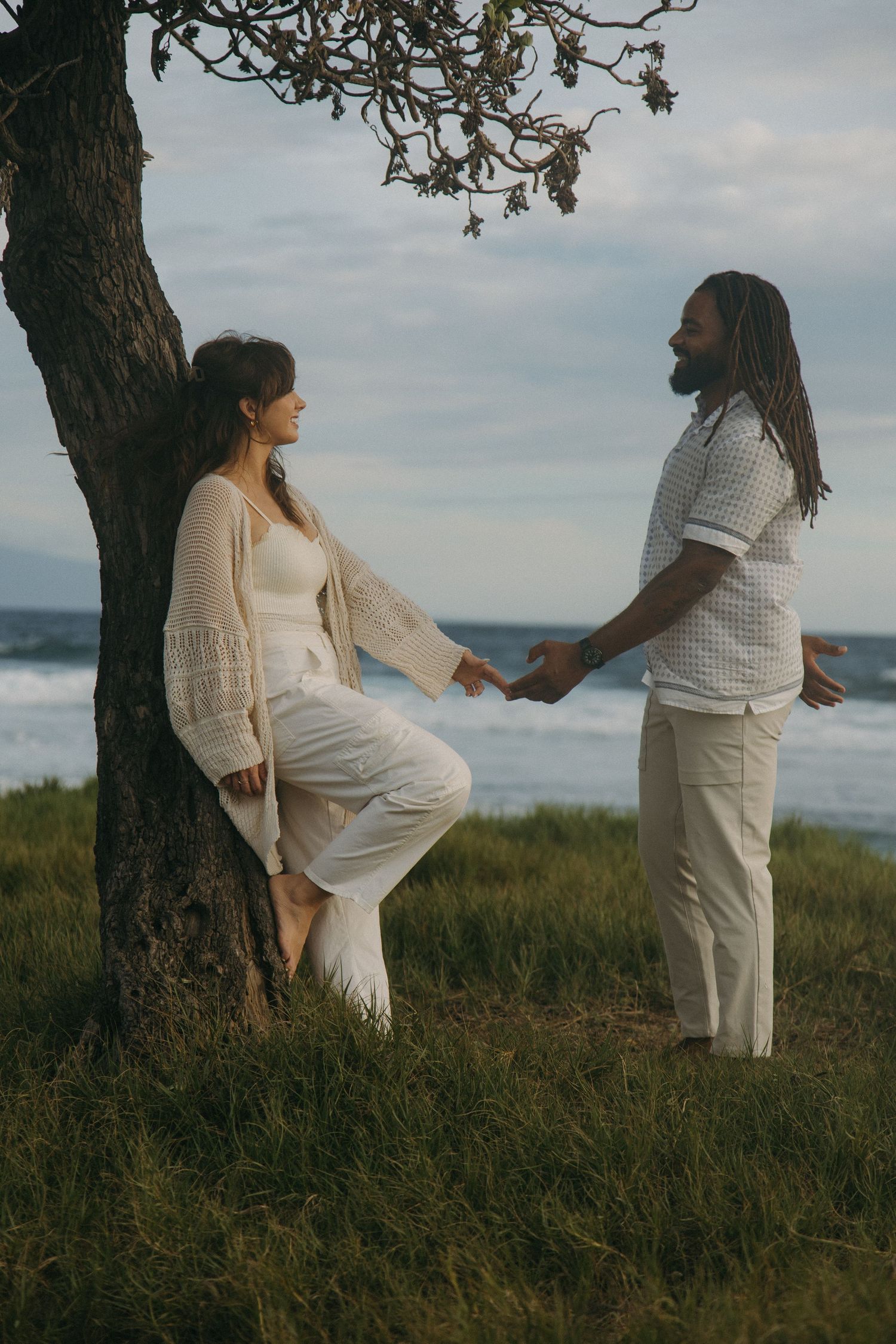 Couple in white outfits holding hands by a tree near the ocean, sharing a romantic moment on a grassy cliff.