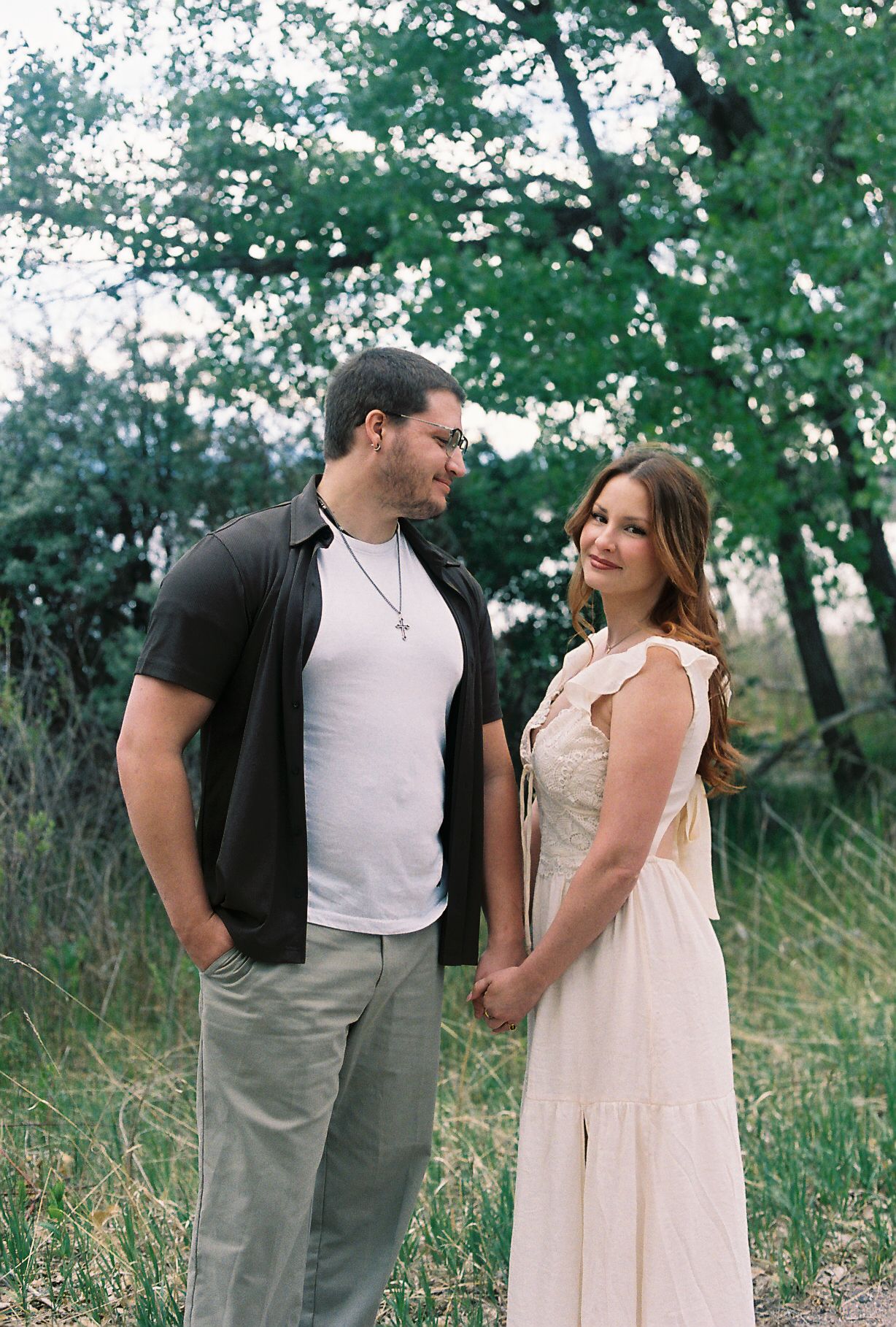 A couple holds hands and gazes at each other in a lush green outdoor setting, the woman in a cream dress and man in casual attire.