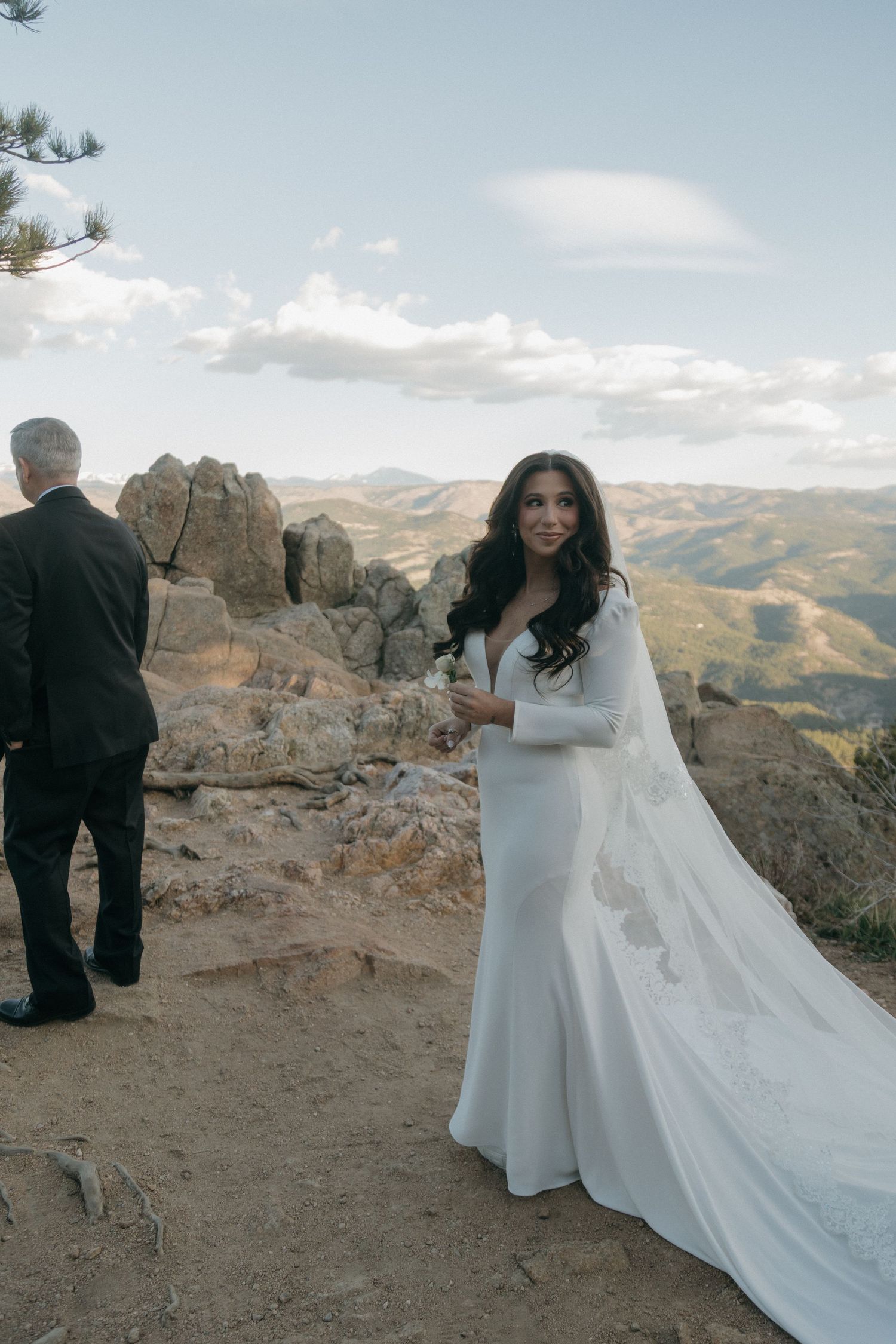 A bride in a white long-sleeve gown with flowing veil smiling at a mountain ceremony.