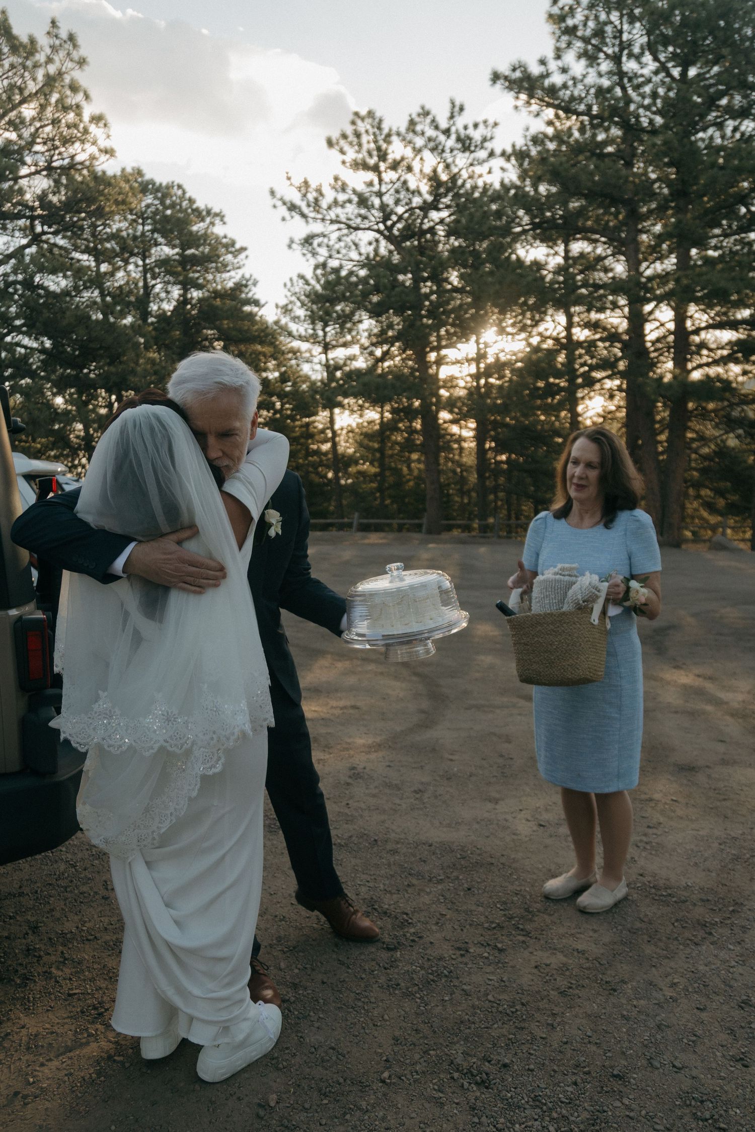 Elderly guest hugs bride in white gown while woman holds wedding cake outdoors.