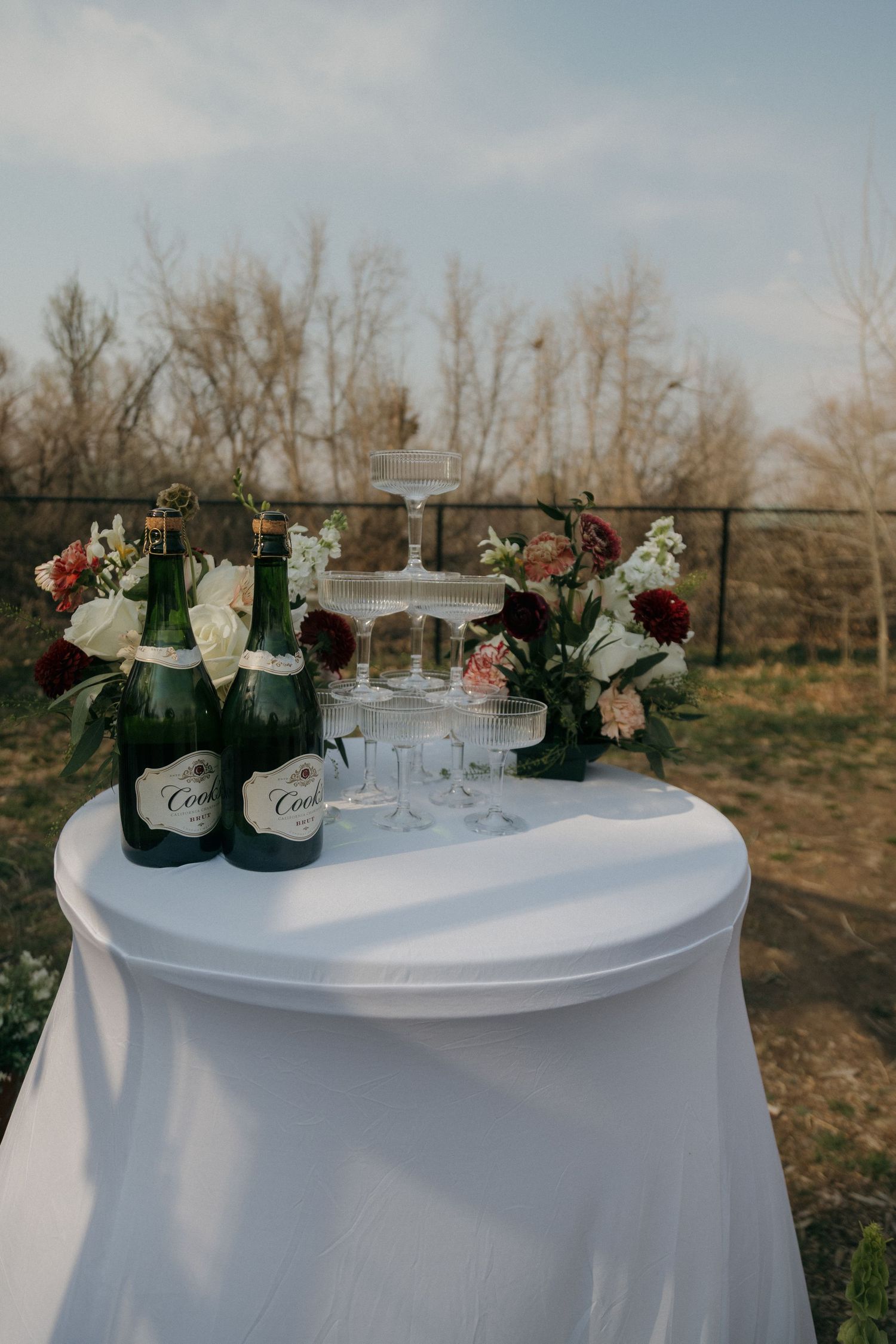 Elegant outdoor wedding champagne tower setup with Cooks bottles and burgundy and white floral arrangements.