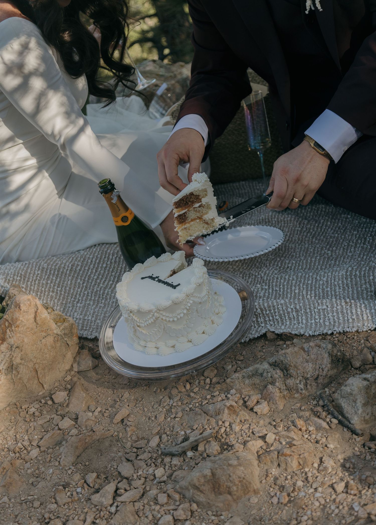 Bride and groom cutting a small white wedding cake outdoors on rocky terrain with a champagne bottle nearby.
