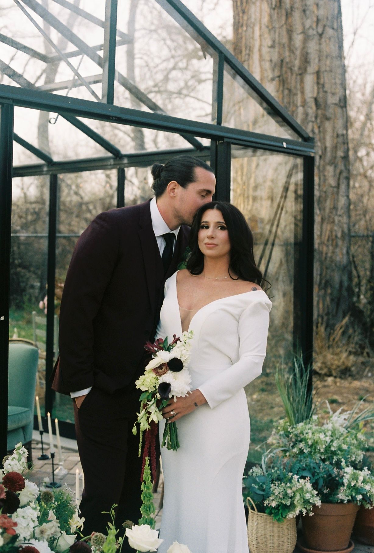 Bride in off-shoulder white gown holds bouquet as groom in burgundy suit kisses her cheek by a greenhouse.