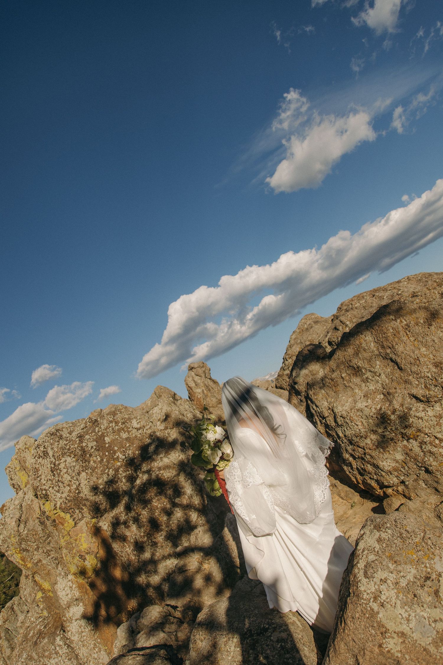 Bride in flowing white wedding dress and veil stands between rocky boulders under blue sky.