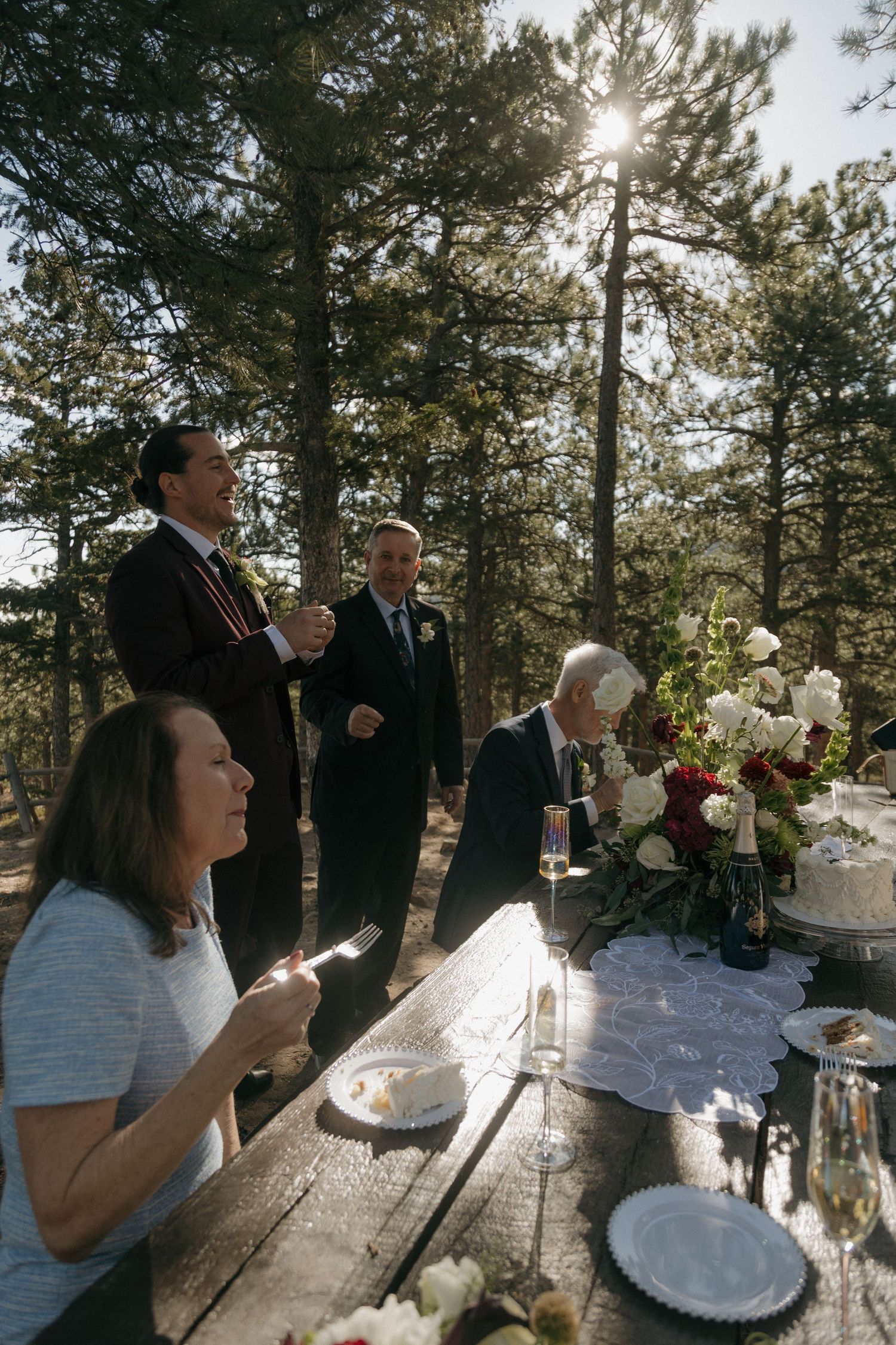 Wedding guests gather around a rustic wooden table with floral arrangements in a sunlit pine forest.