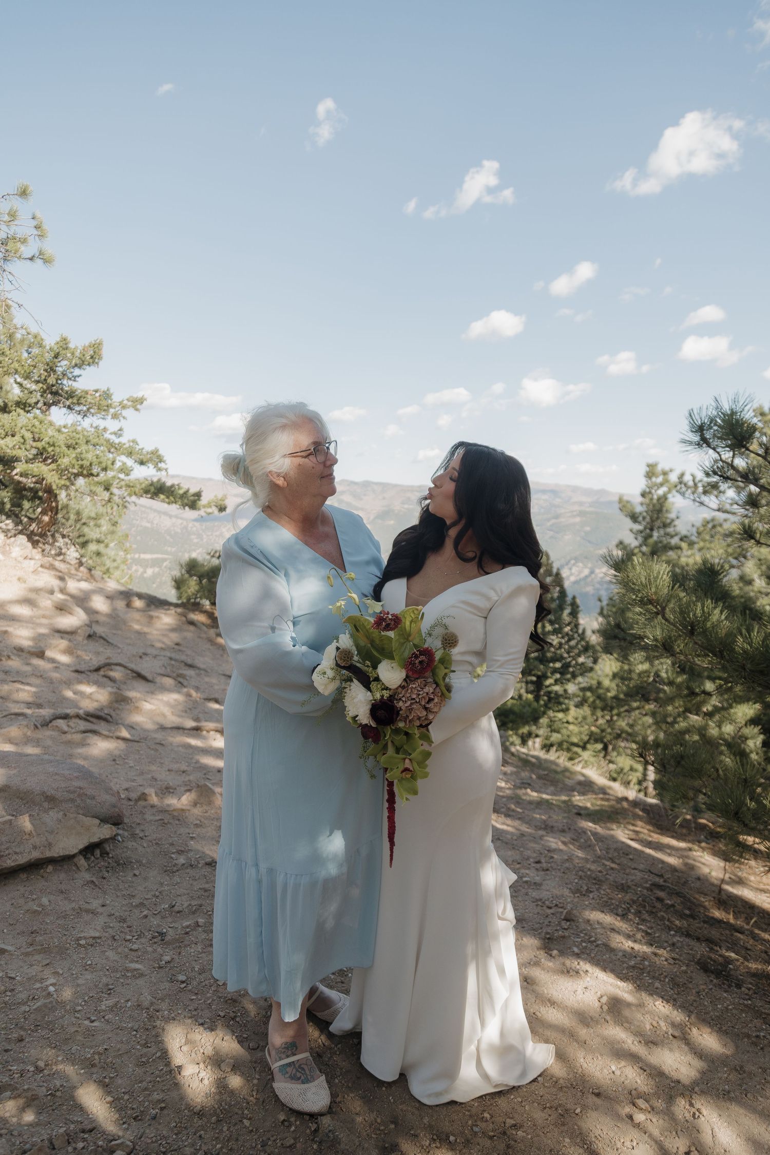 Bride in white gown sharing tender moment with elderly grandmother at outdoor mountain wedding ceremony.