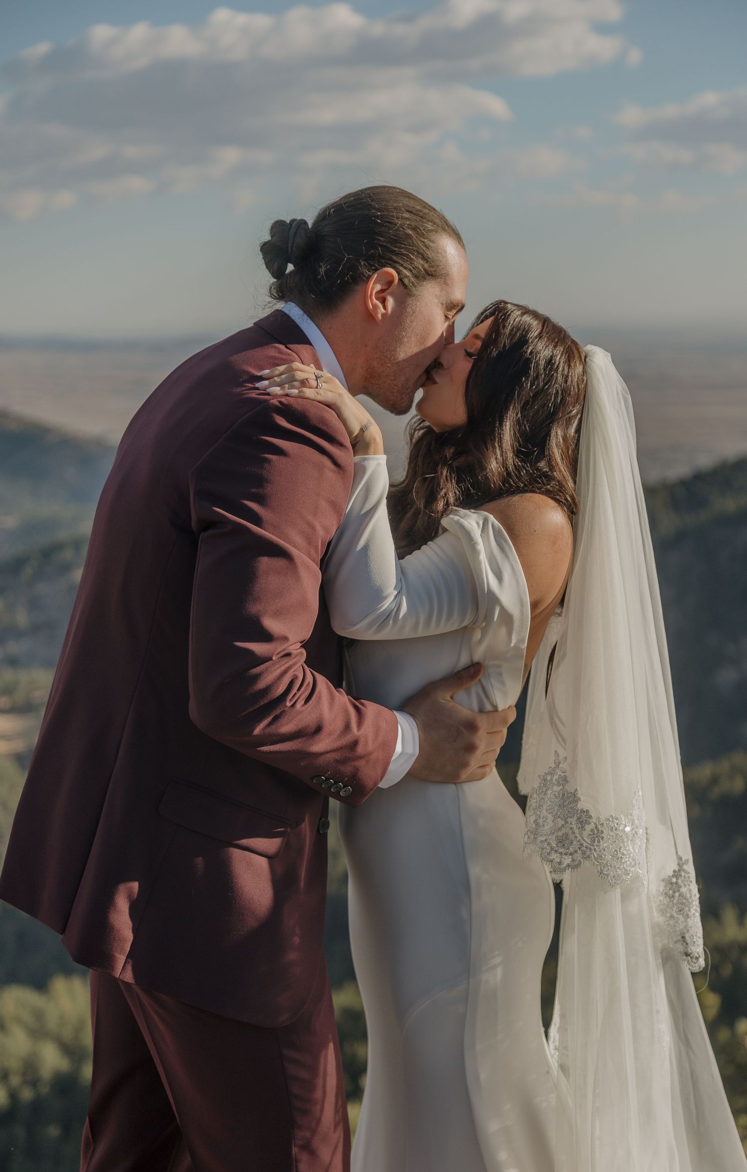 Bride and groom share a first kiss in wedding attire with a scenic mountain valley backdrop.