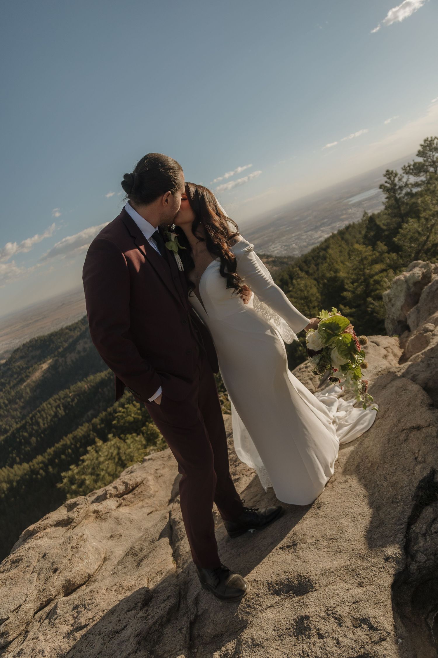 Bride and groom share a kiss on a rocky mountain summit with panoramic views below.