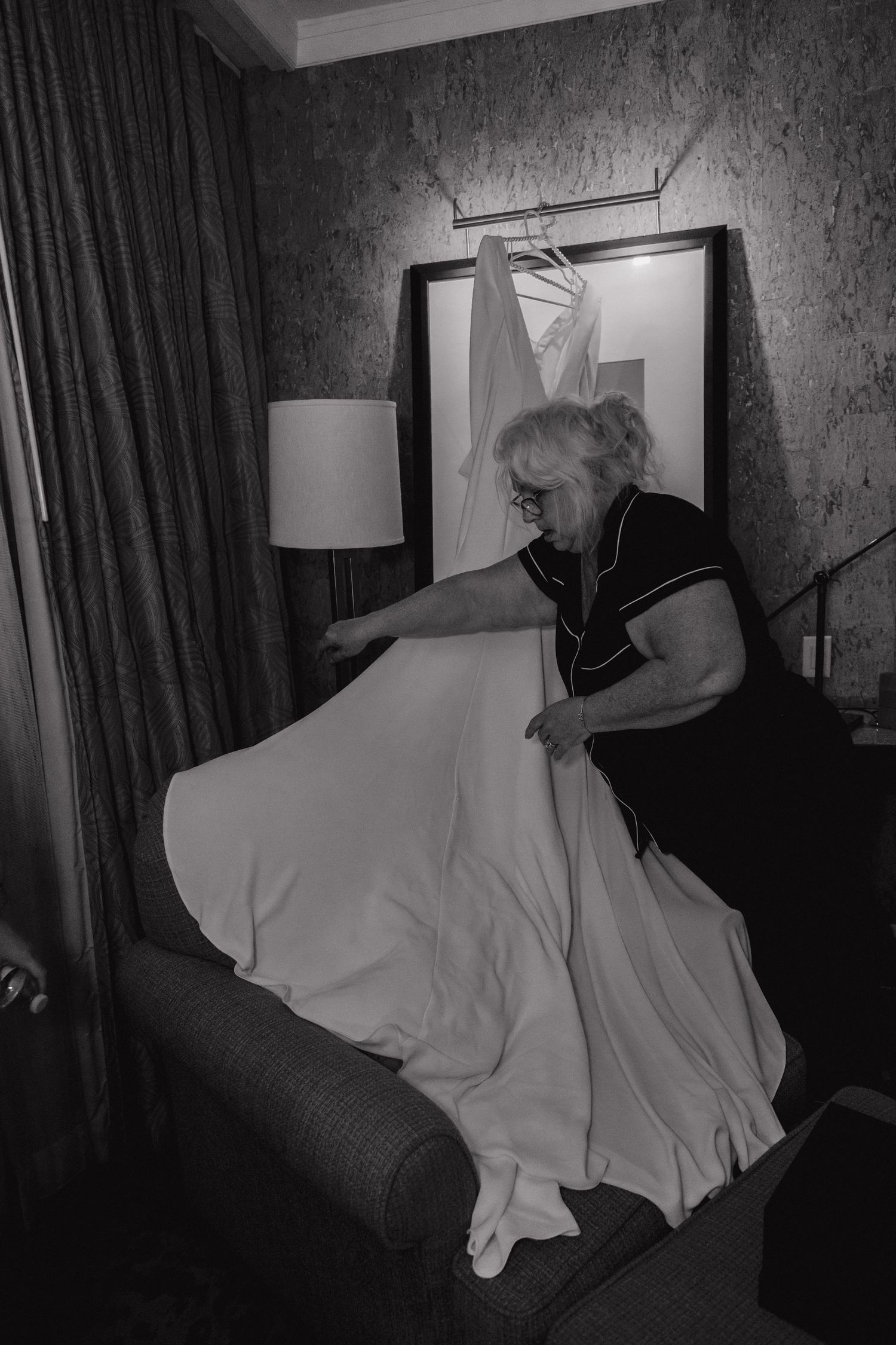 Older woman in black dress arranging a white wedding gown in a hotel room.