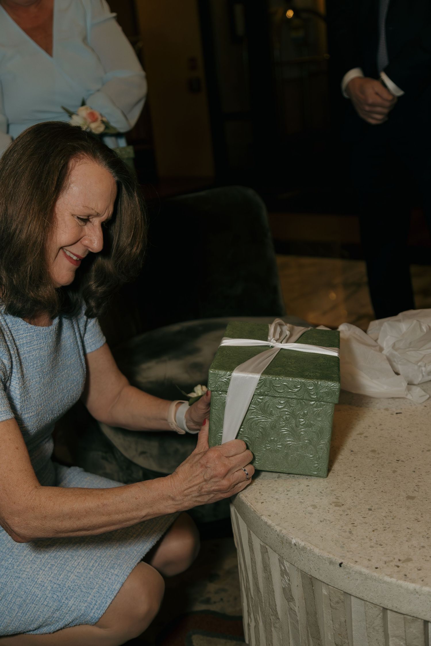 Smiling woman in blue dress opening a green gift box with white ribbon at a wedding celebration.