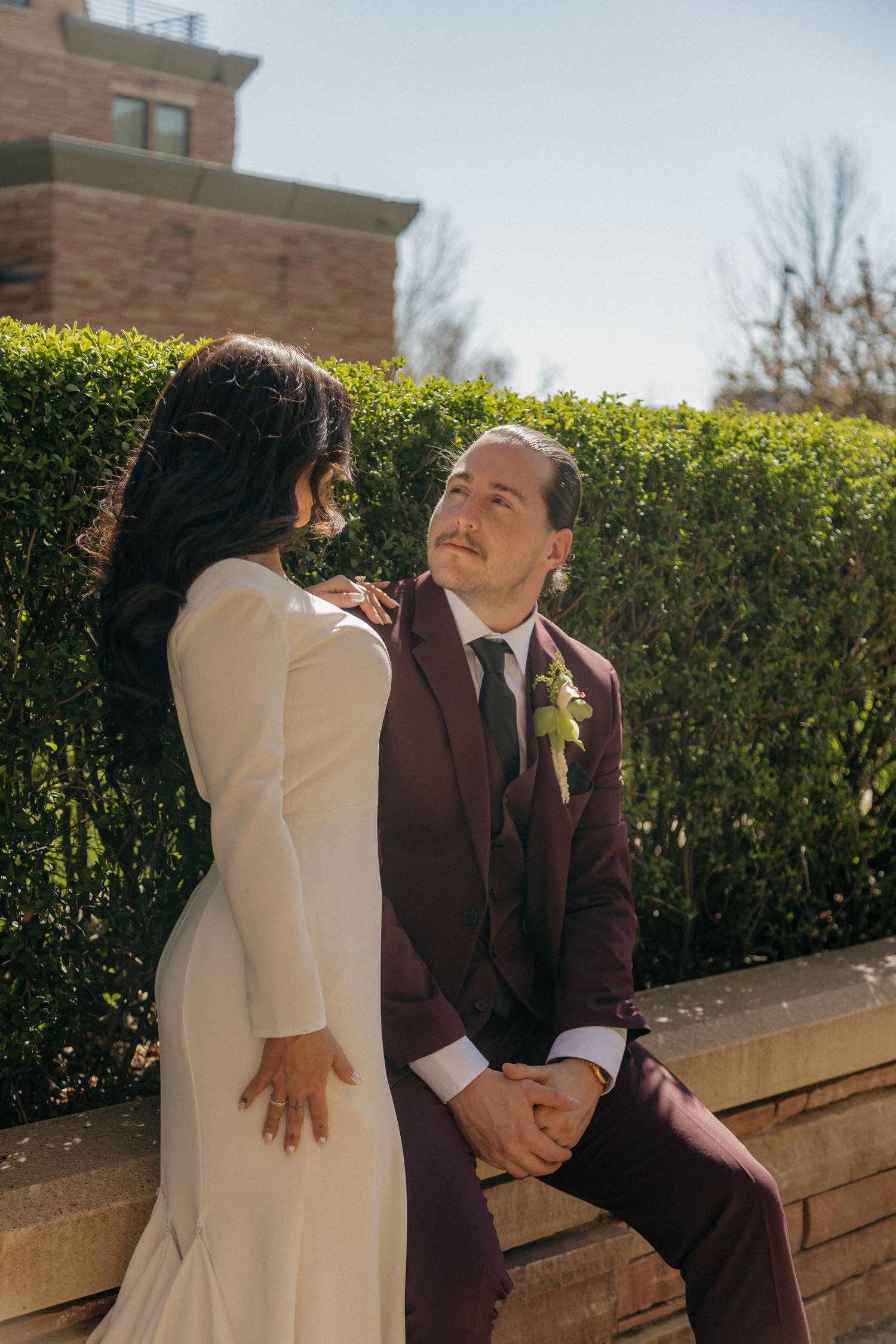 Bride and groom in burgundy suit sitting on stone ledge surrounded by green hedges.