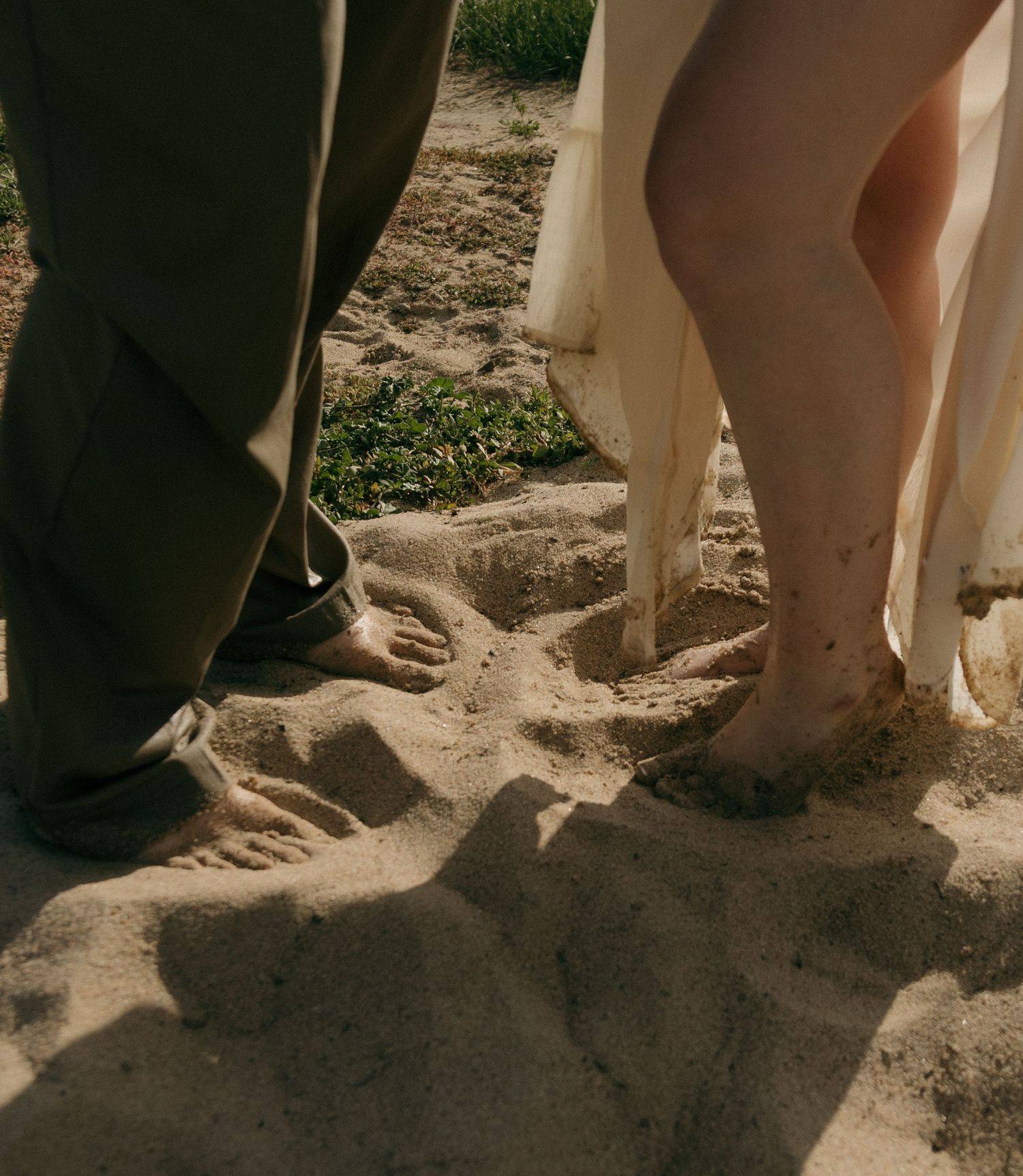 Close-up of a barefoot couple standing together in sandy soil, womans white dress hem visible beside mans khaki pants.