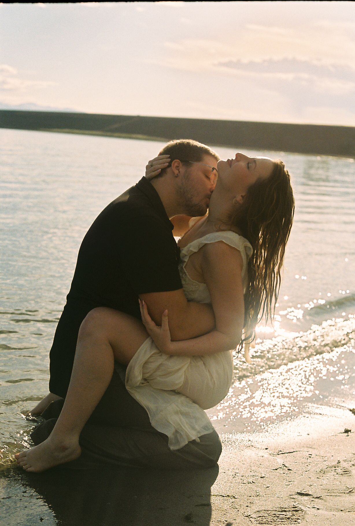 Couple sharing a romantic kiss while sitting at the water's edge in golden sunlight.