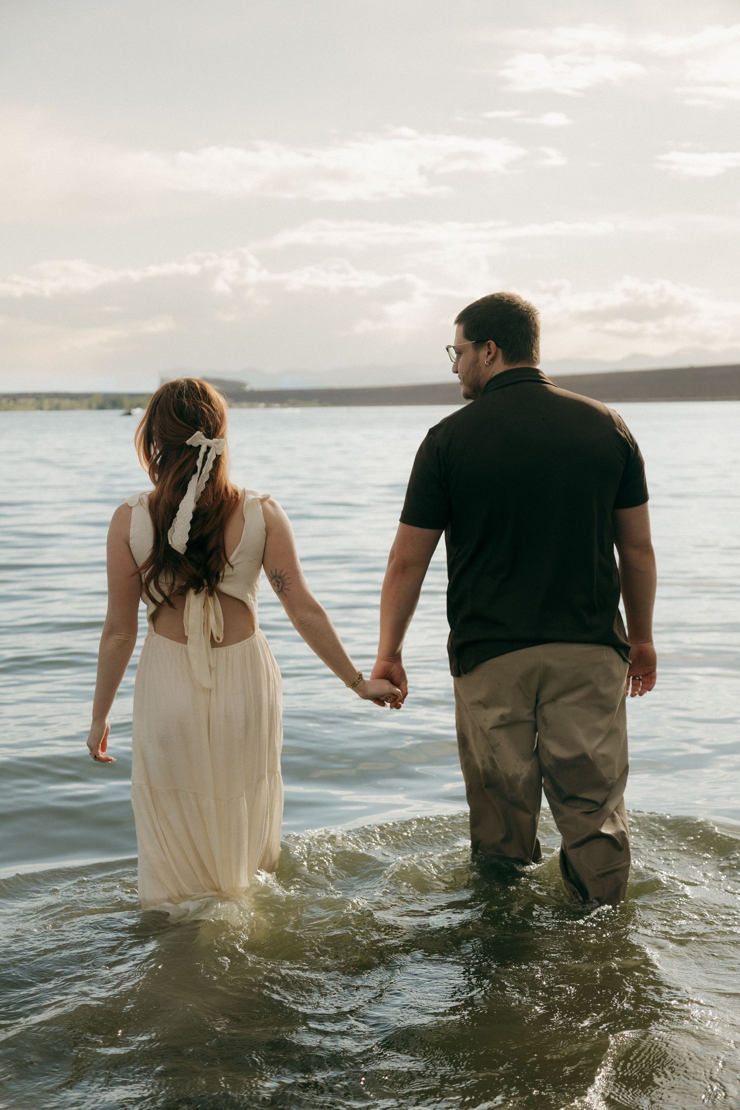 Couple holding hands wading into a calm lake at dusk, woman in white dress with bow.