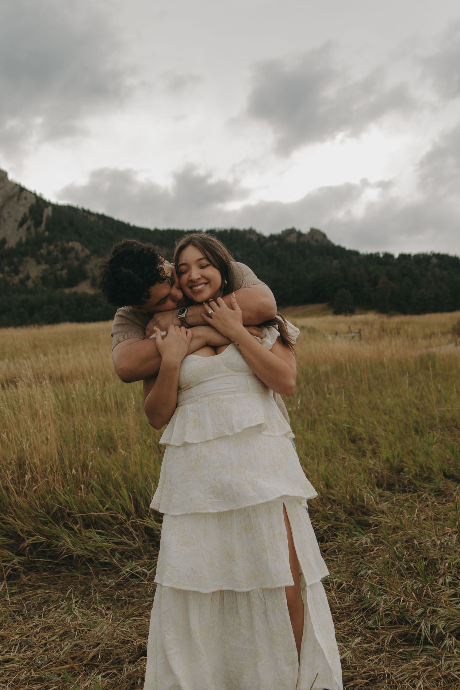 Man embracing woman in white ruffled dress from behind, both laughing in a rustic mountain meadow.