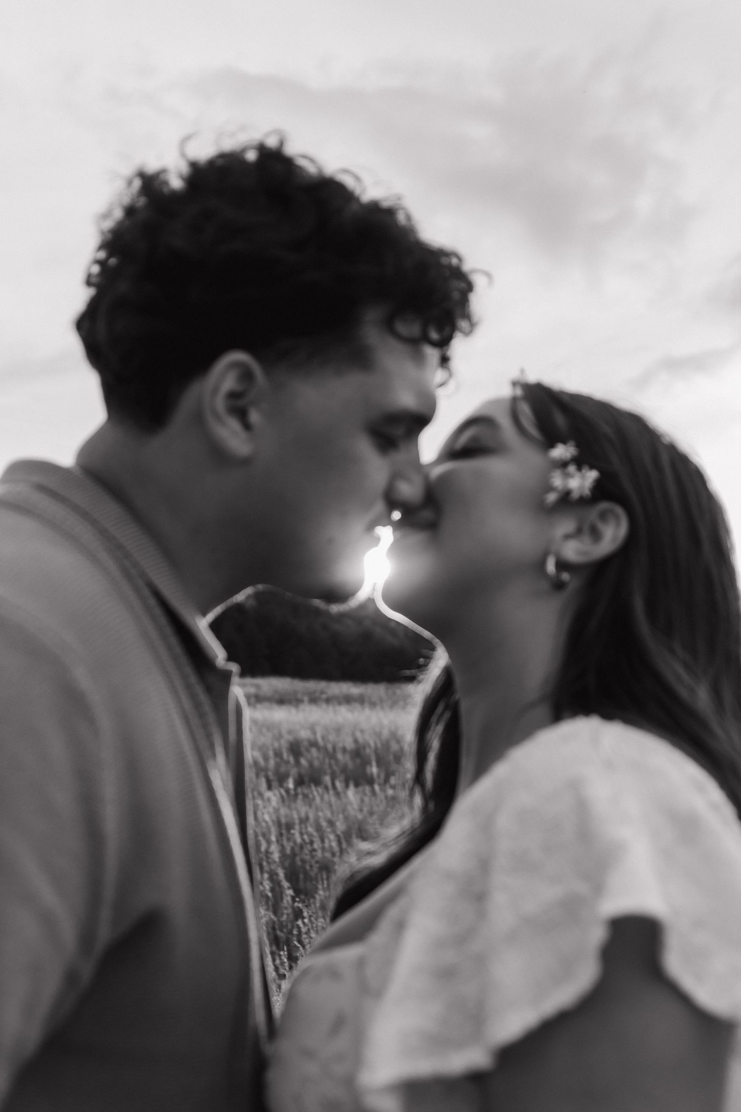 Black and white photo of a couple sharing a romantic kiss in a field at sunset.