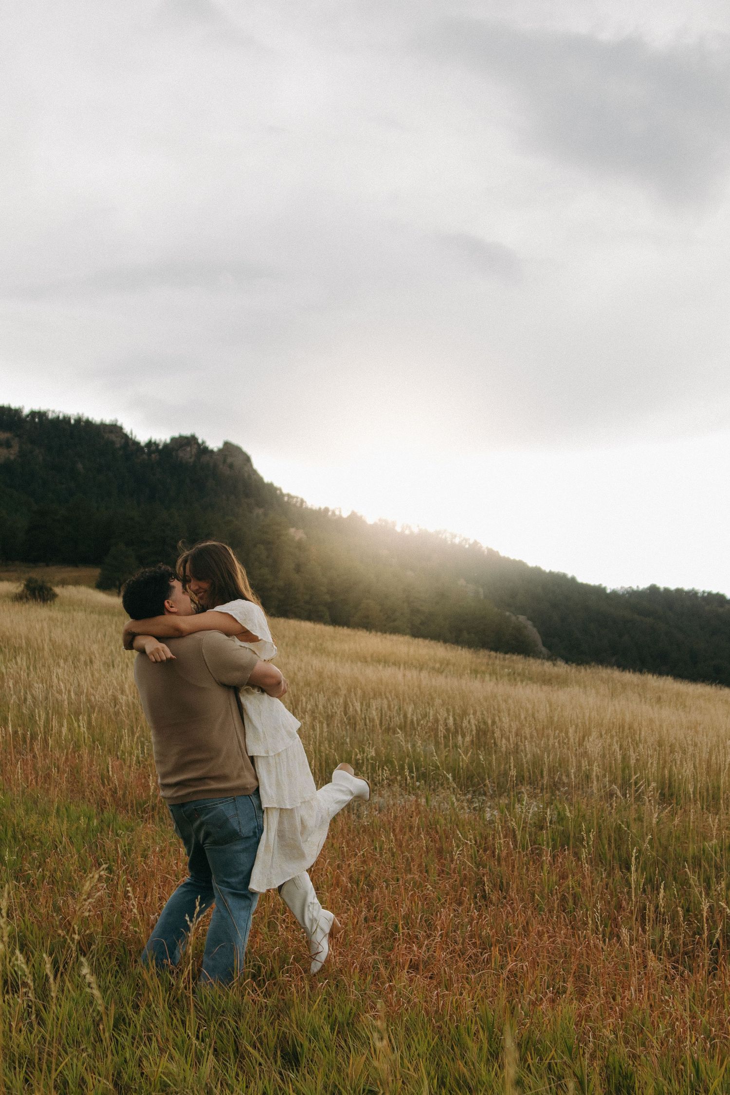 Couple embracing and kissing in a golden meadow with mountain forest backdrop during sunset.