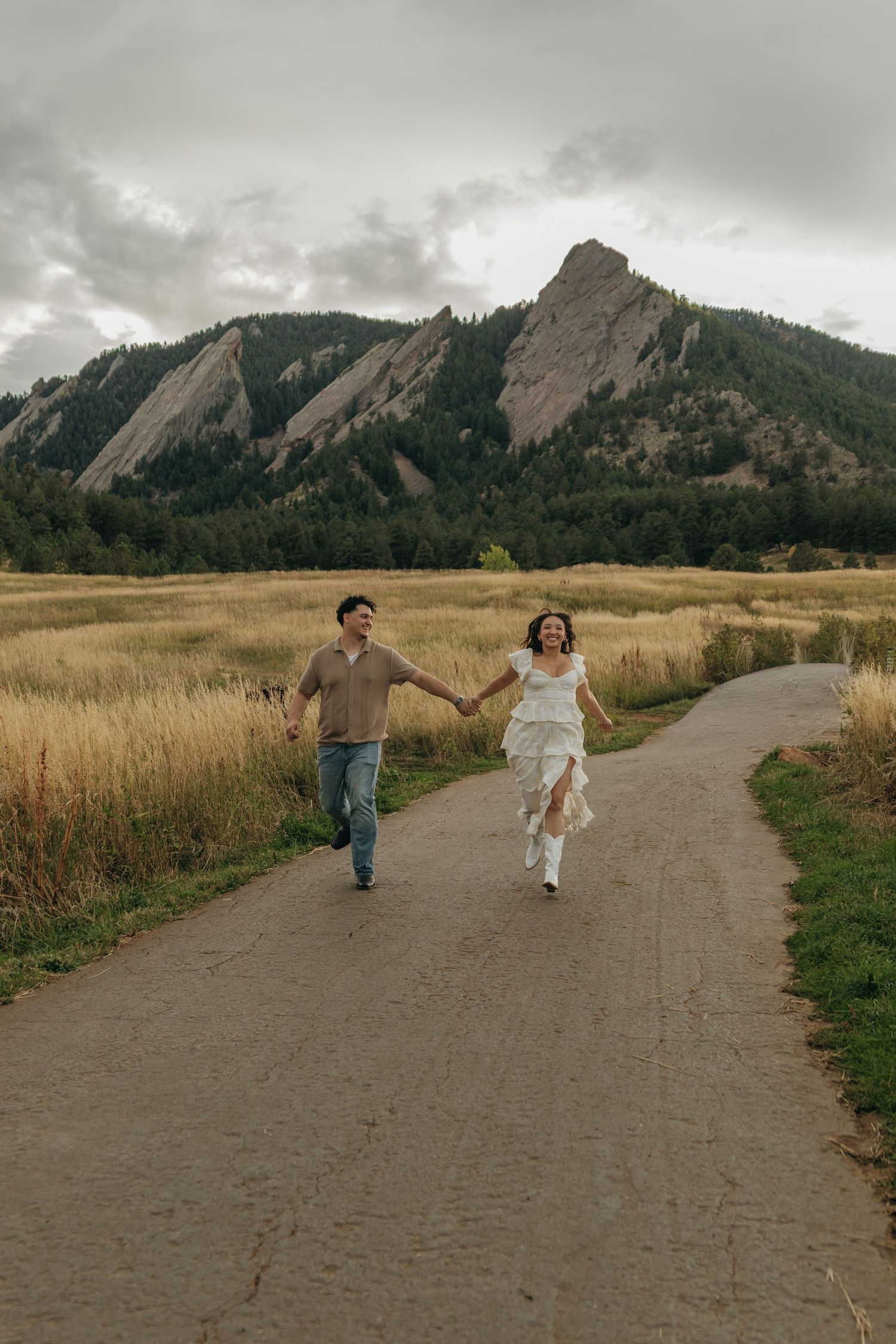 Couple holding hands running down path with dramatic Flatiron mountains in background.