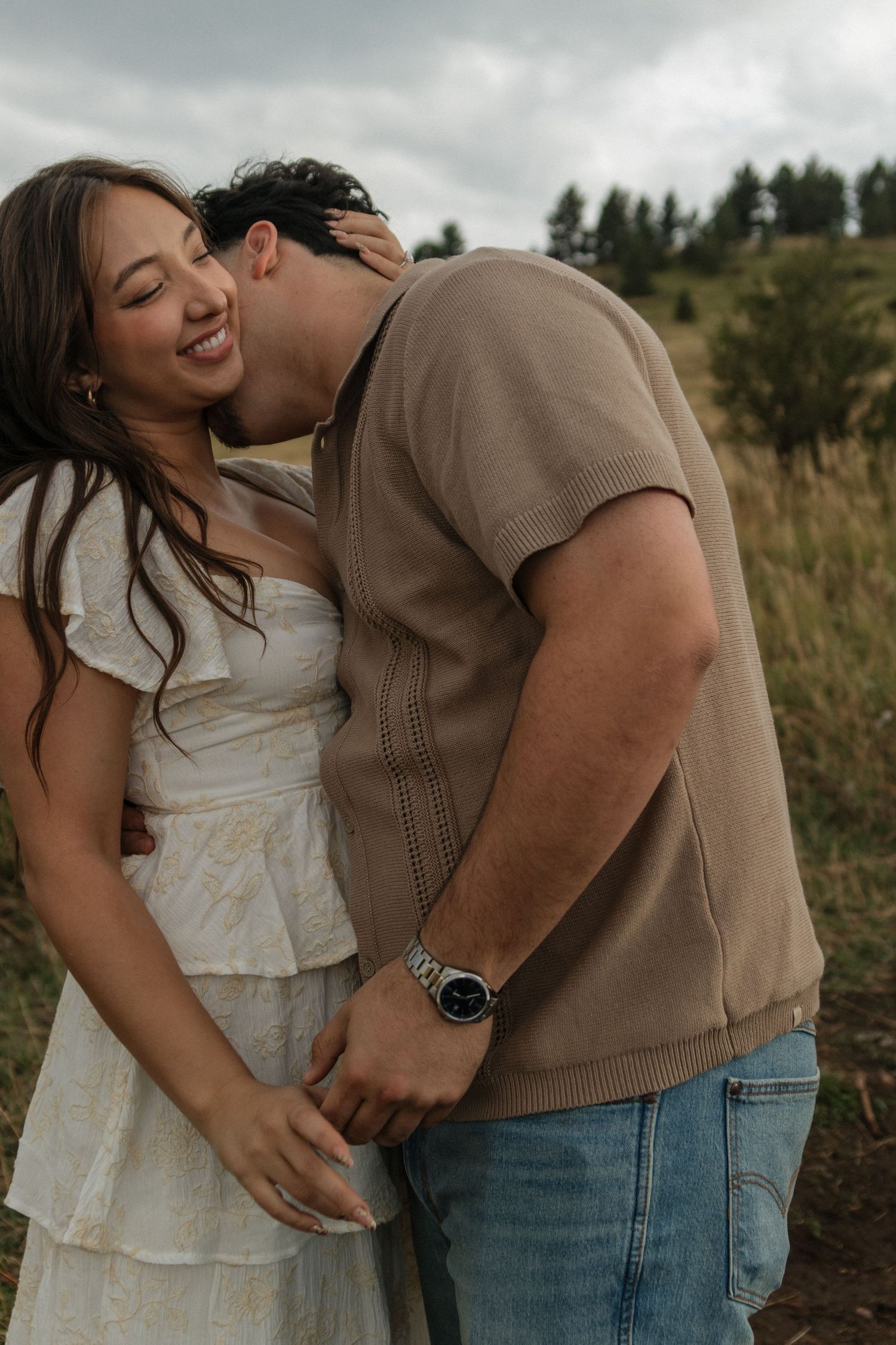 Man kissing woman on the cheek as she smiles in a grassy mountain field during an engagement session.