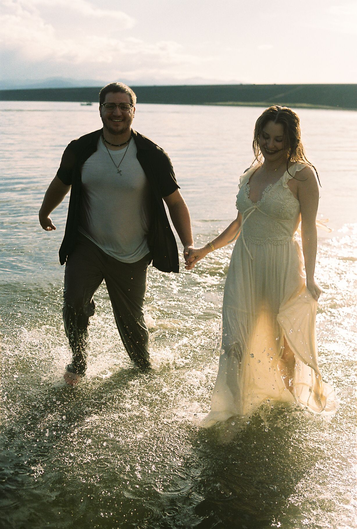 Happy couple holding hands and walking through shallow lake water during a sunny outdoor engagement session.