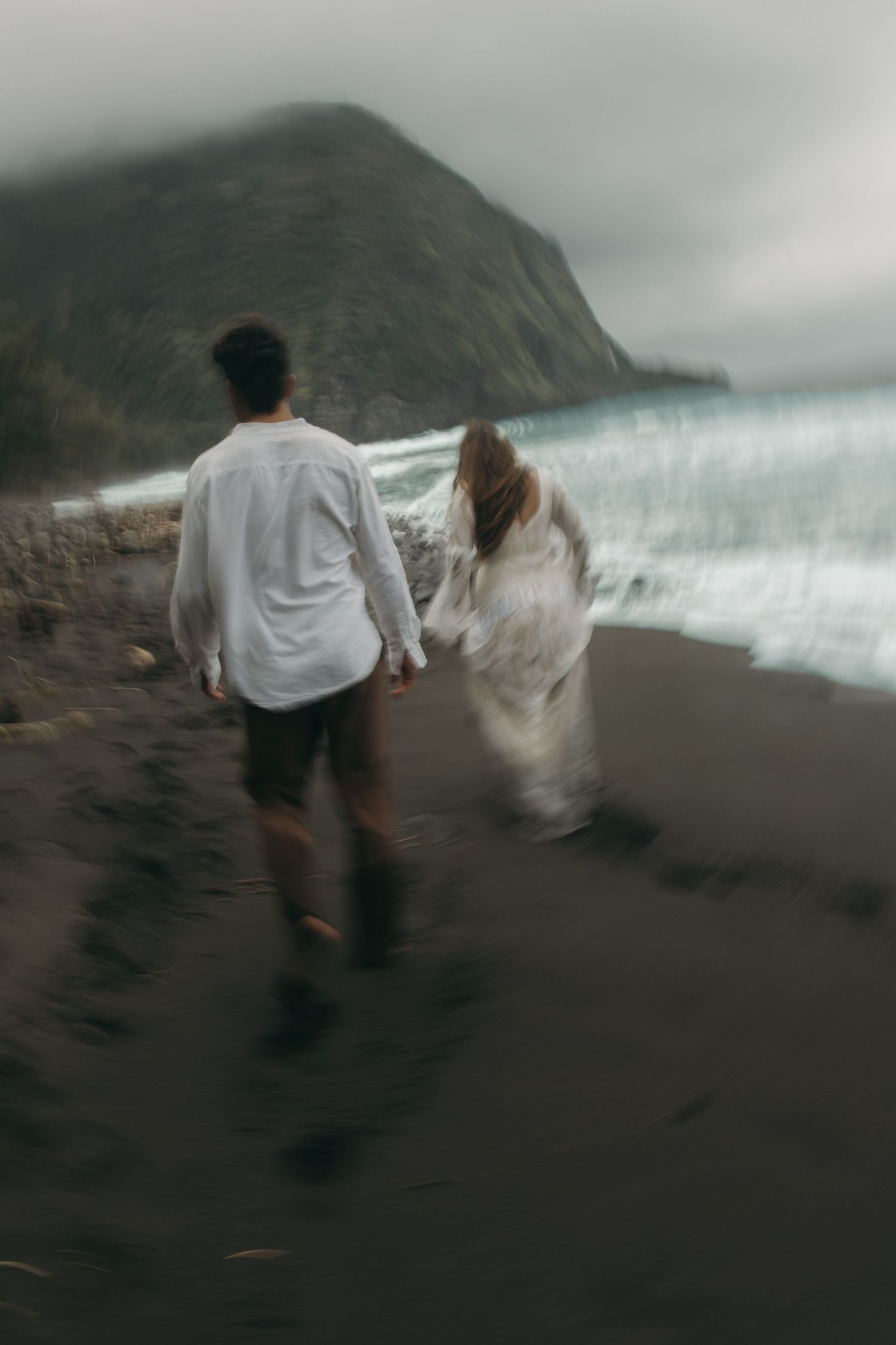 Blurred motion photo of couple walking hand in hand on dark sand beach with misty mountain backdrop.