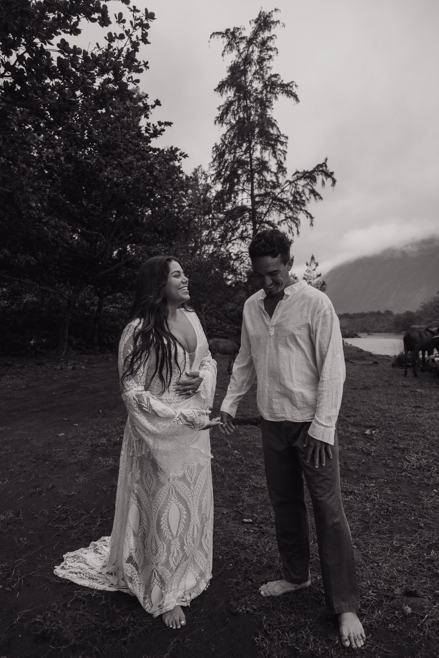 Black and white photo of smiling bride in lace gown holding hands with groom barefoot outdoors.