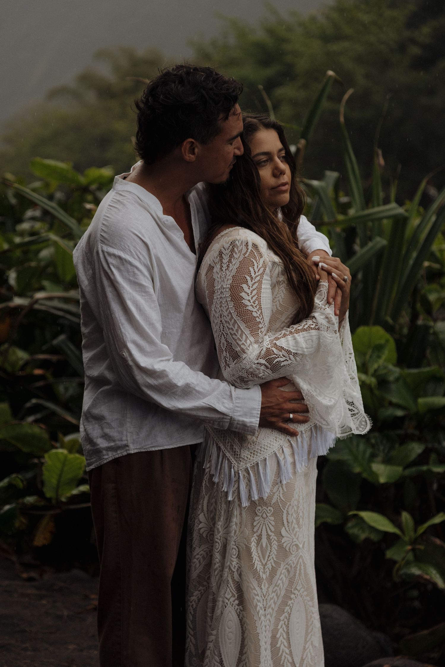 Groom embraces bride in a lace boho wedding dress among lush tropical greenery in soft light.