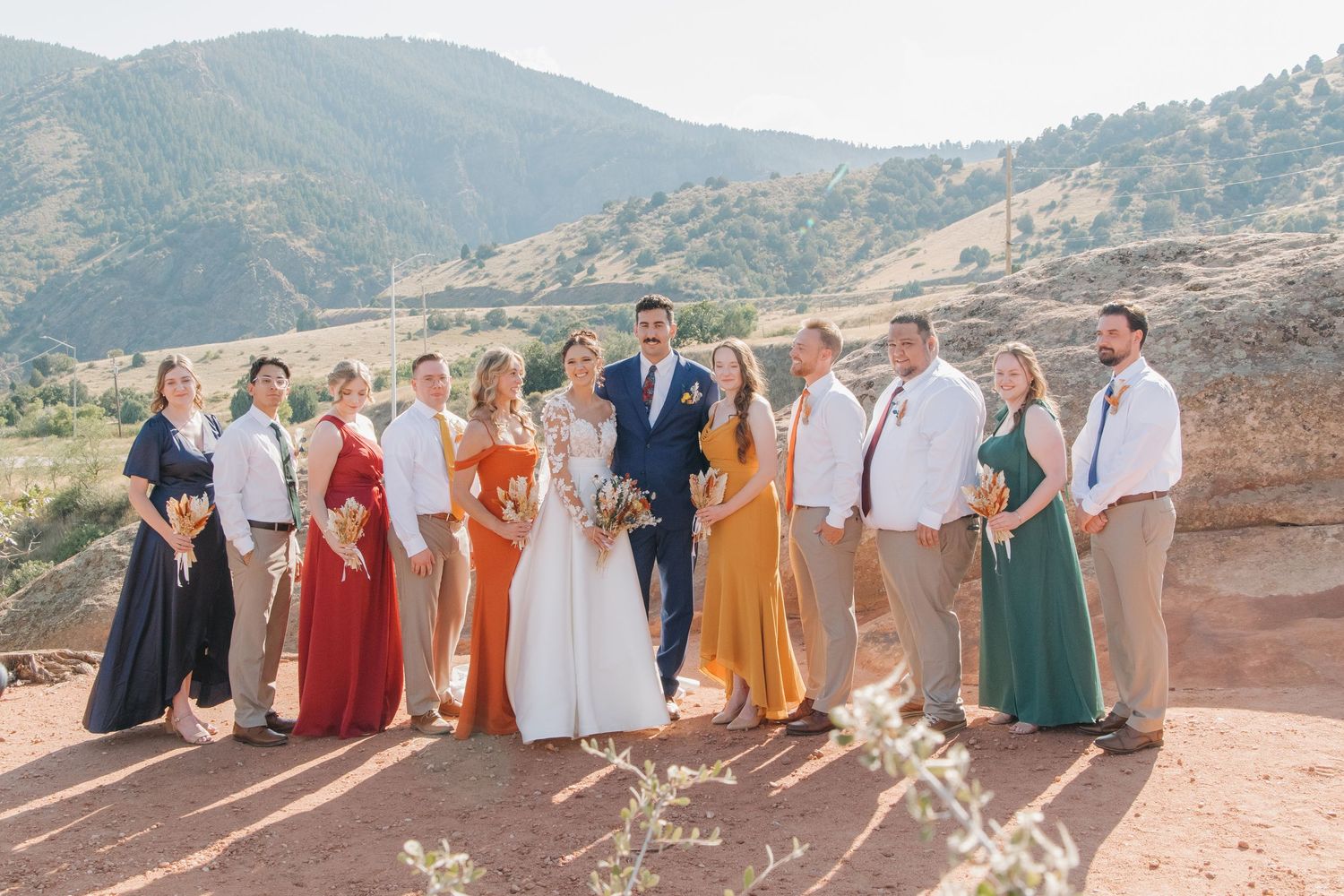 Wedding party members in coordinated attire stand lined up against a mountain landscape.
