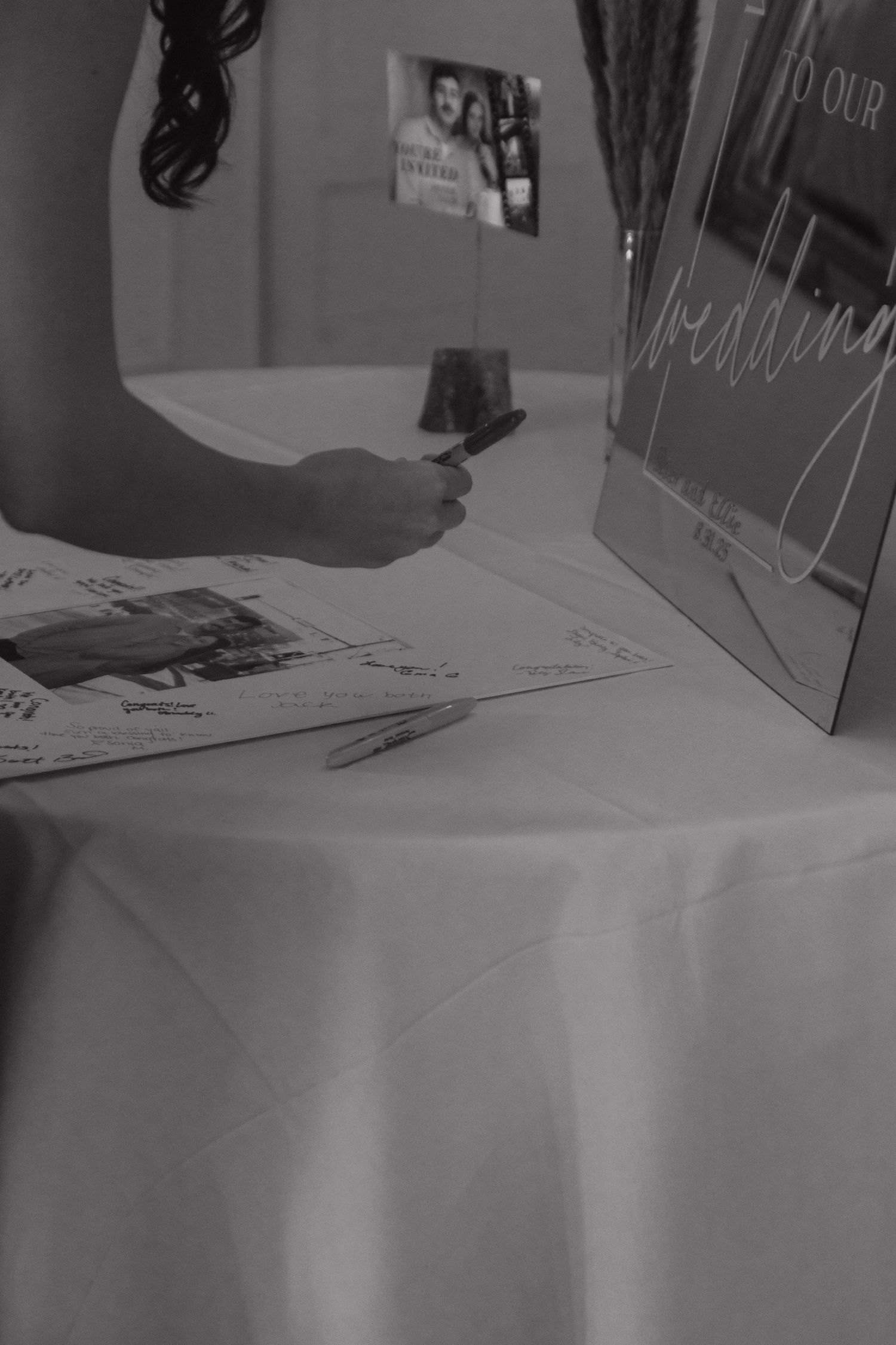 Black and white image of someone writing in a wedding guestbook on a white tablecloth.